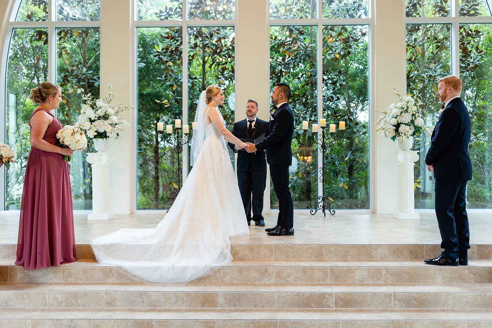 A wedding ceremony inside a building with tall windows and greenery outside. The bride and groom are holding hands facing each other, with an officiant behind them. To the left, a bridesmaid in a mauve dress holds a bouquet, and to the right, a groom