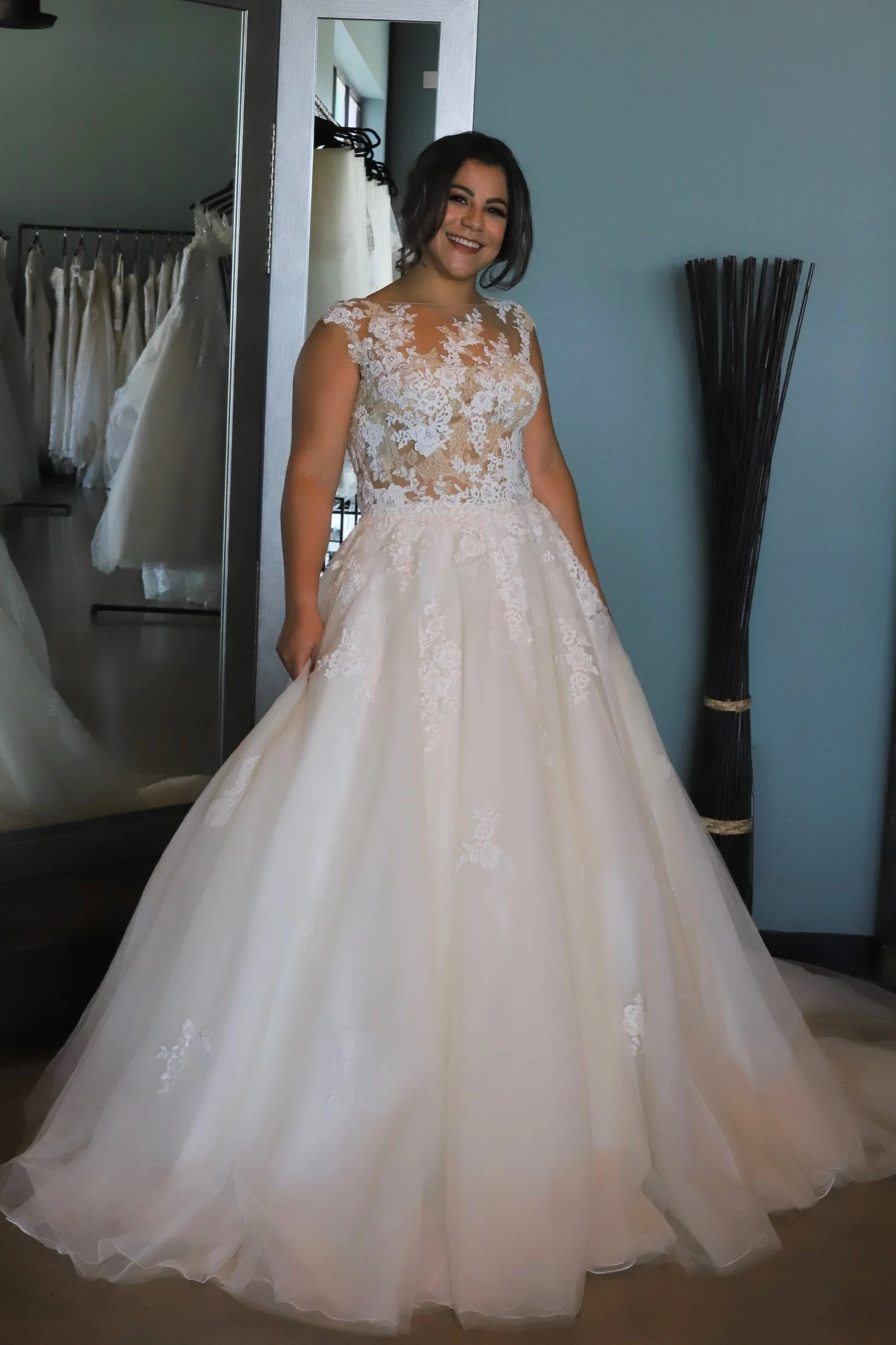 Woman trying on a wedding dress with a lace top and full skirt, smiling in a bridal boutique fitting room.