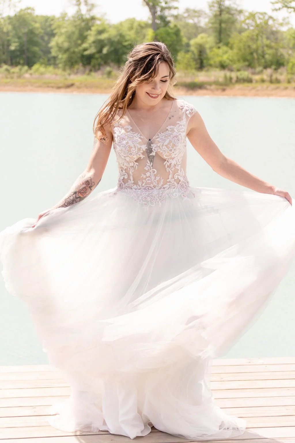 A woman wearing a wedding dress with lace details, holding out the skirt, standing on a wooden dock near a body of water with trees in the background.