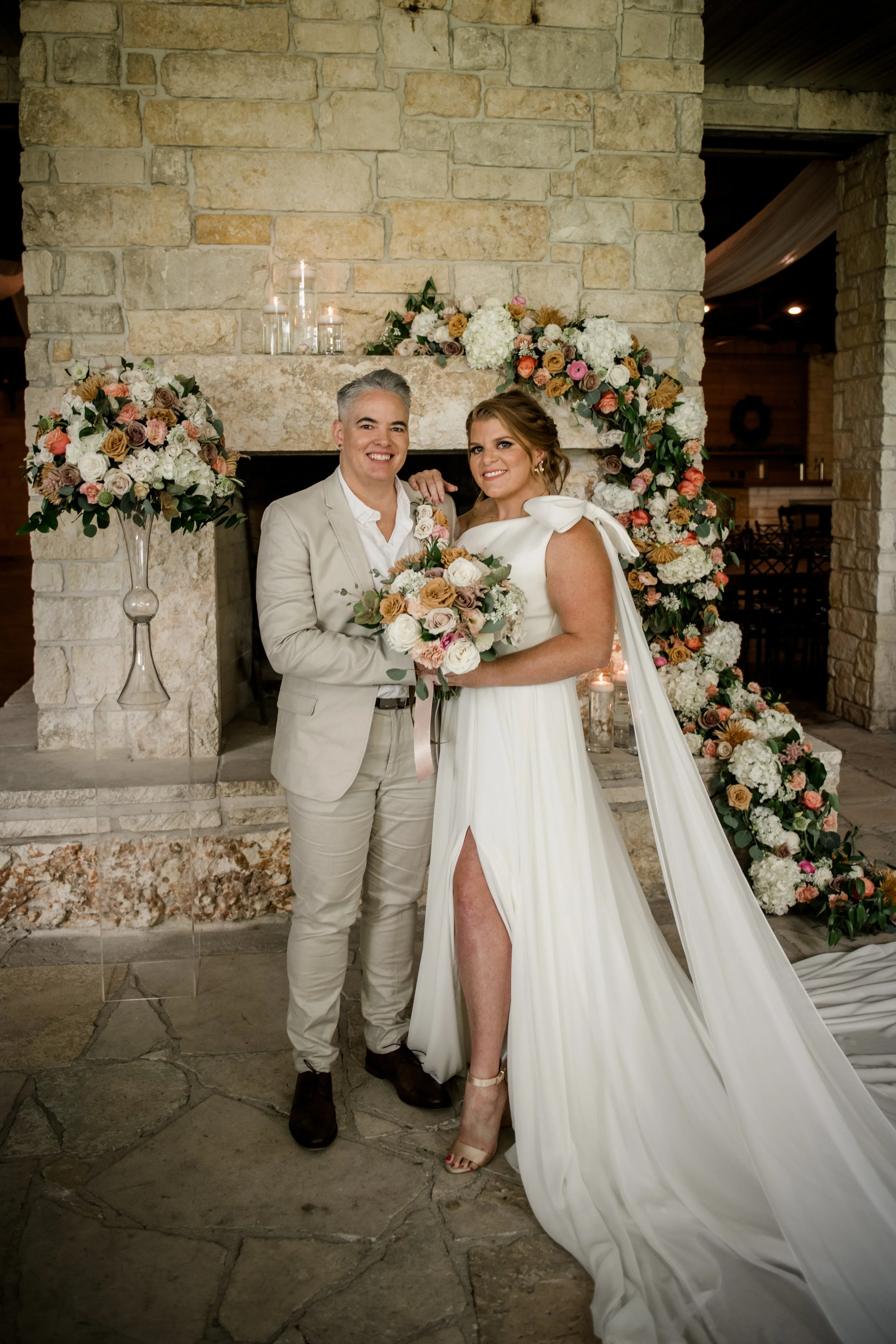 Two women in wedding attire standing in front of a stone fireplace decorated with floral arrangements and candles, posing for a photo at a wedding.