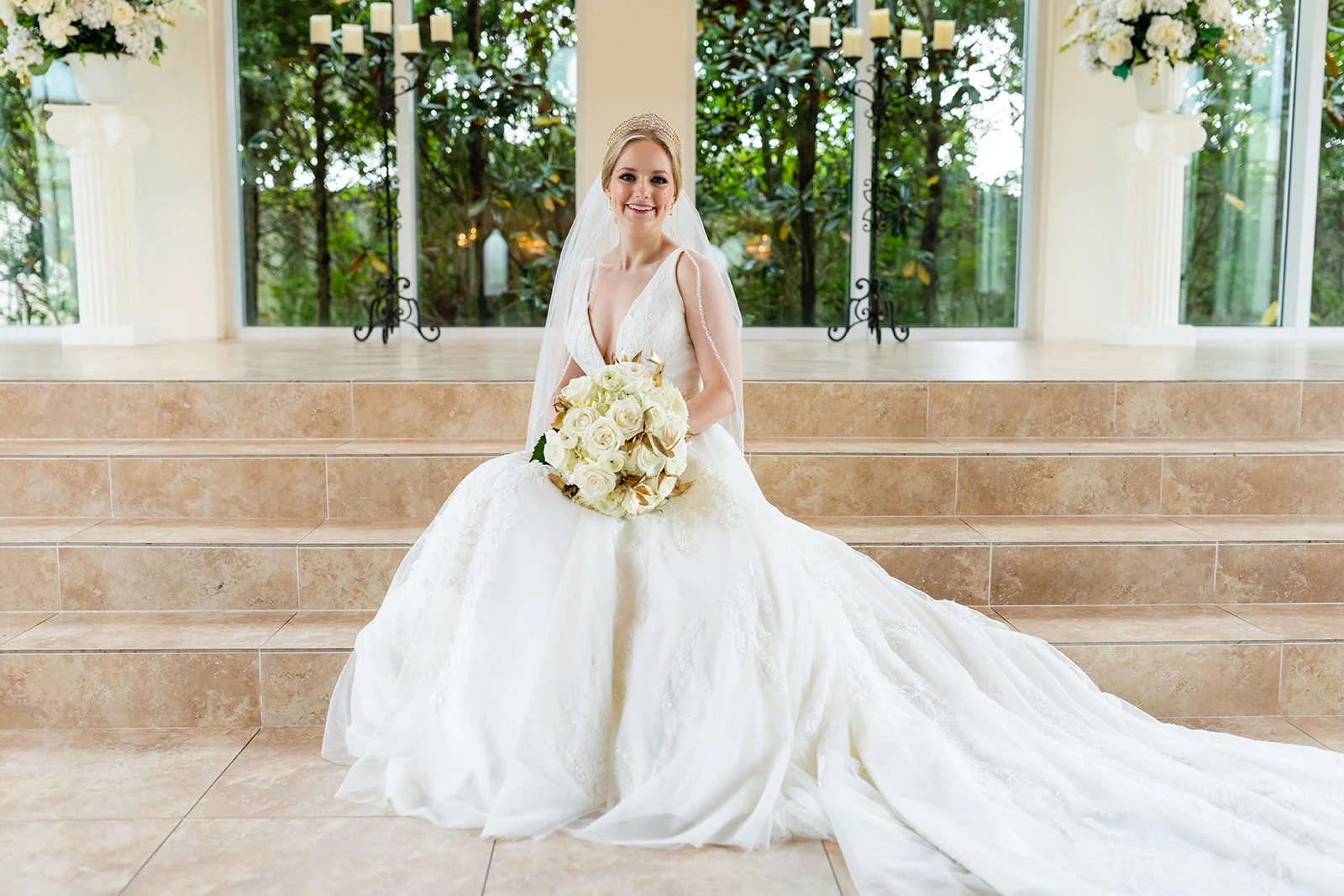 Bride in a white wedding dress sitting on stairs, holding a bouquet of white roses, with large windows and greenery in the background.