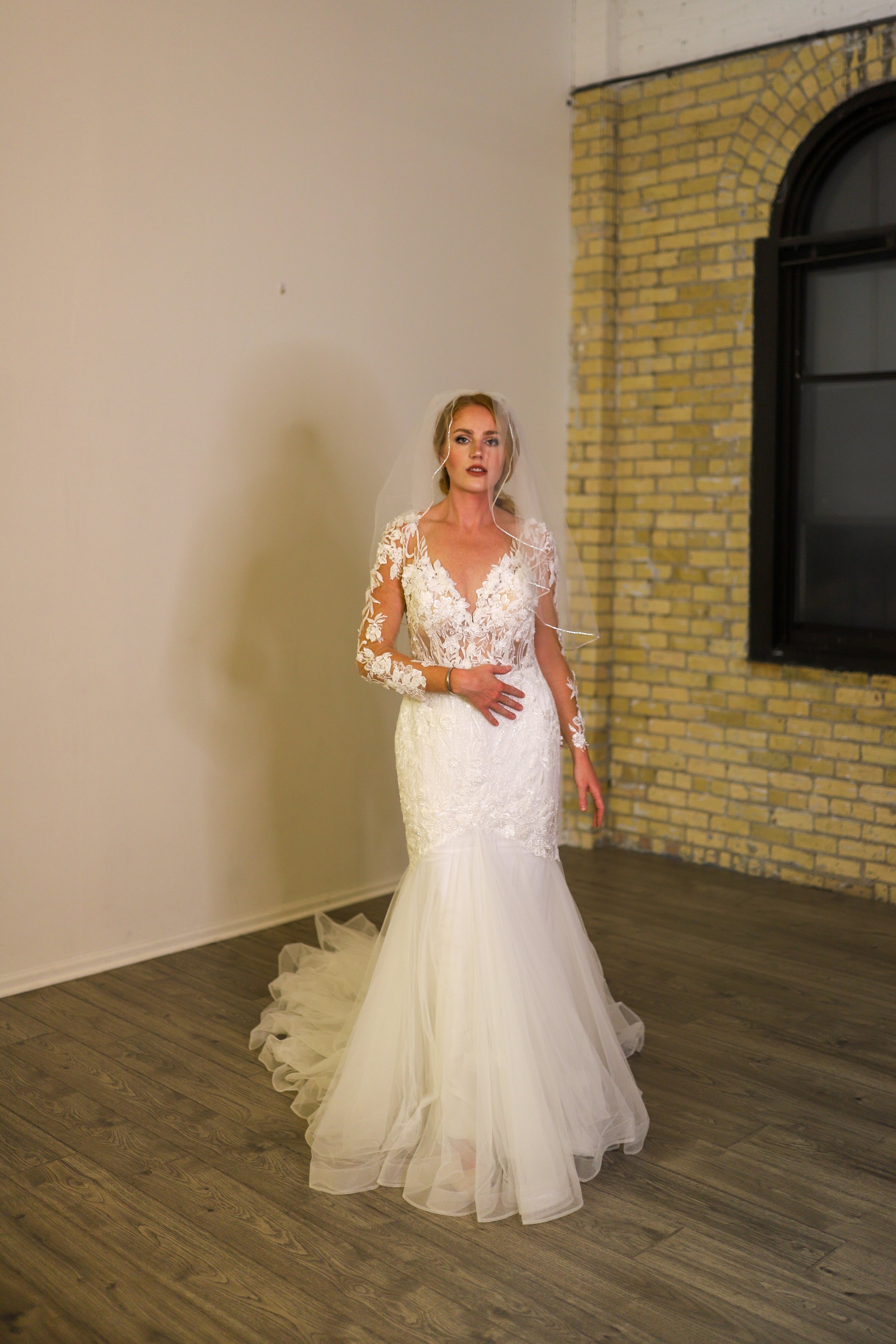 A woman in a white lace wedding dress standing in a room with wooden floors, a brick wall, and a large black window.
