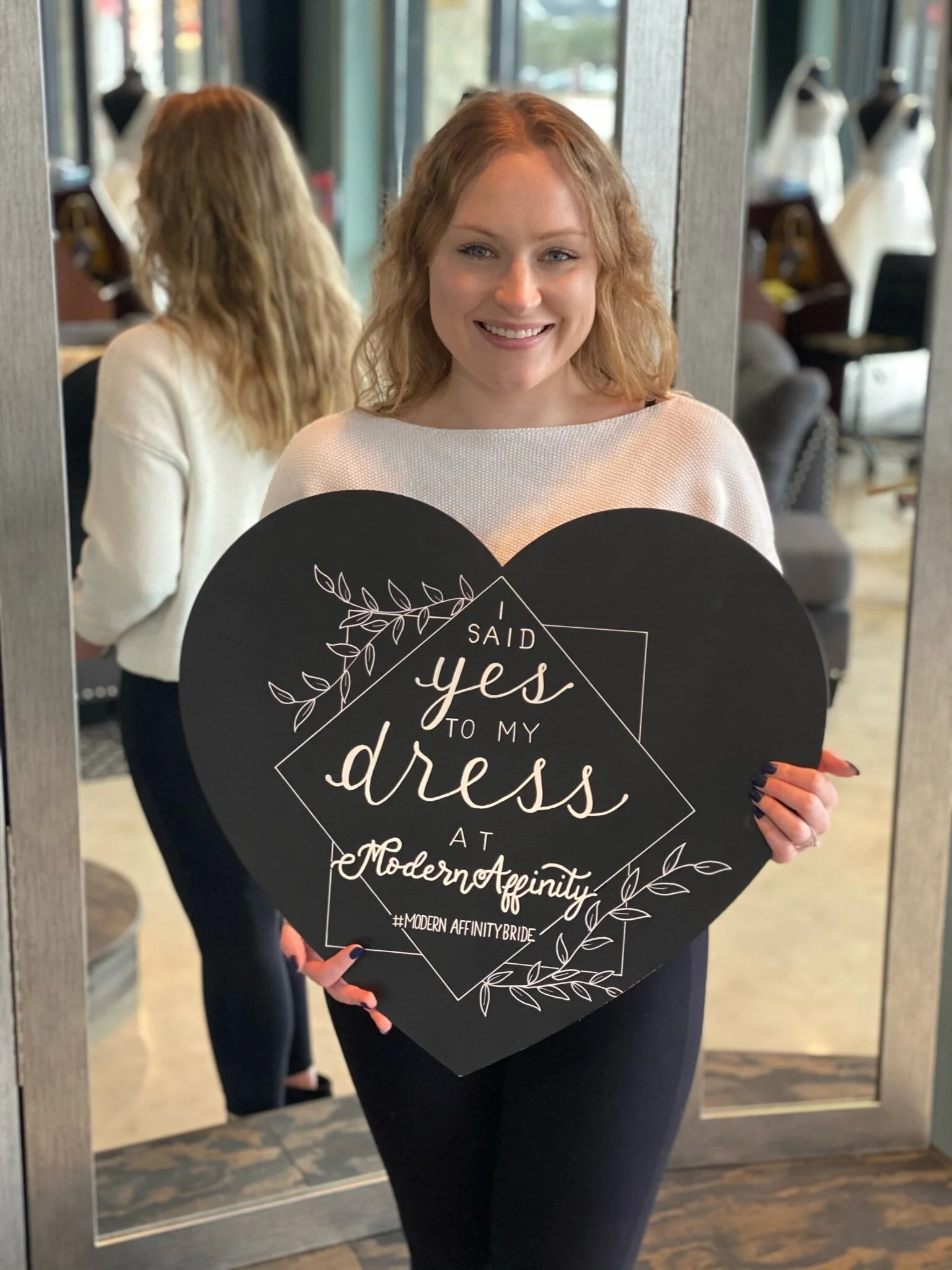 A woman smiling in front of a mirror holding a black heart-shaped sign that reads 'I said yes to my dress at Modern Affinity'.