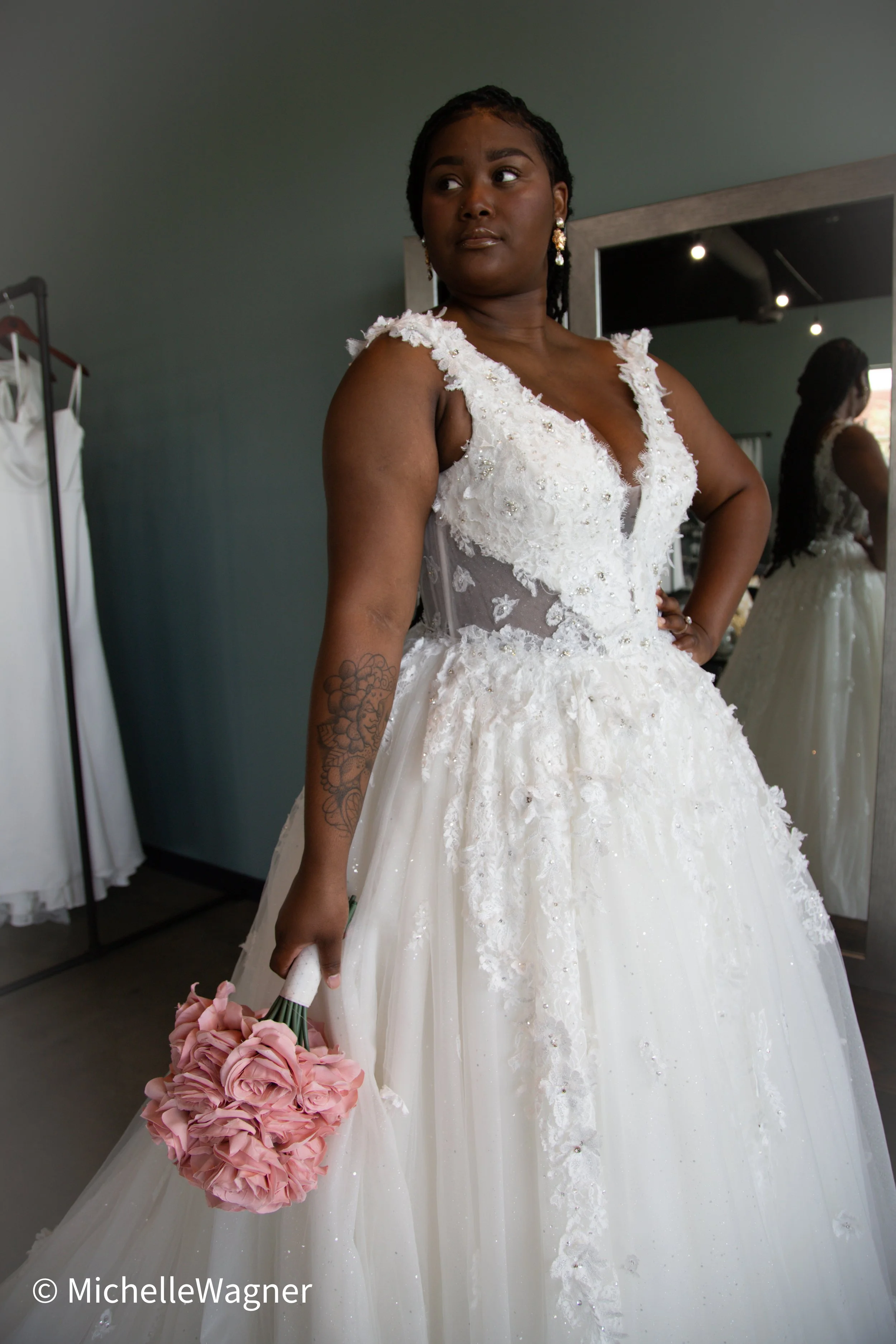Woman in a white wedding dress holding a pink bouquet standing in front of a mirror in a fitting room.
