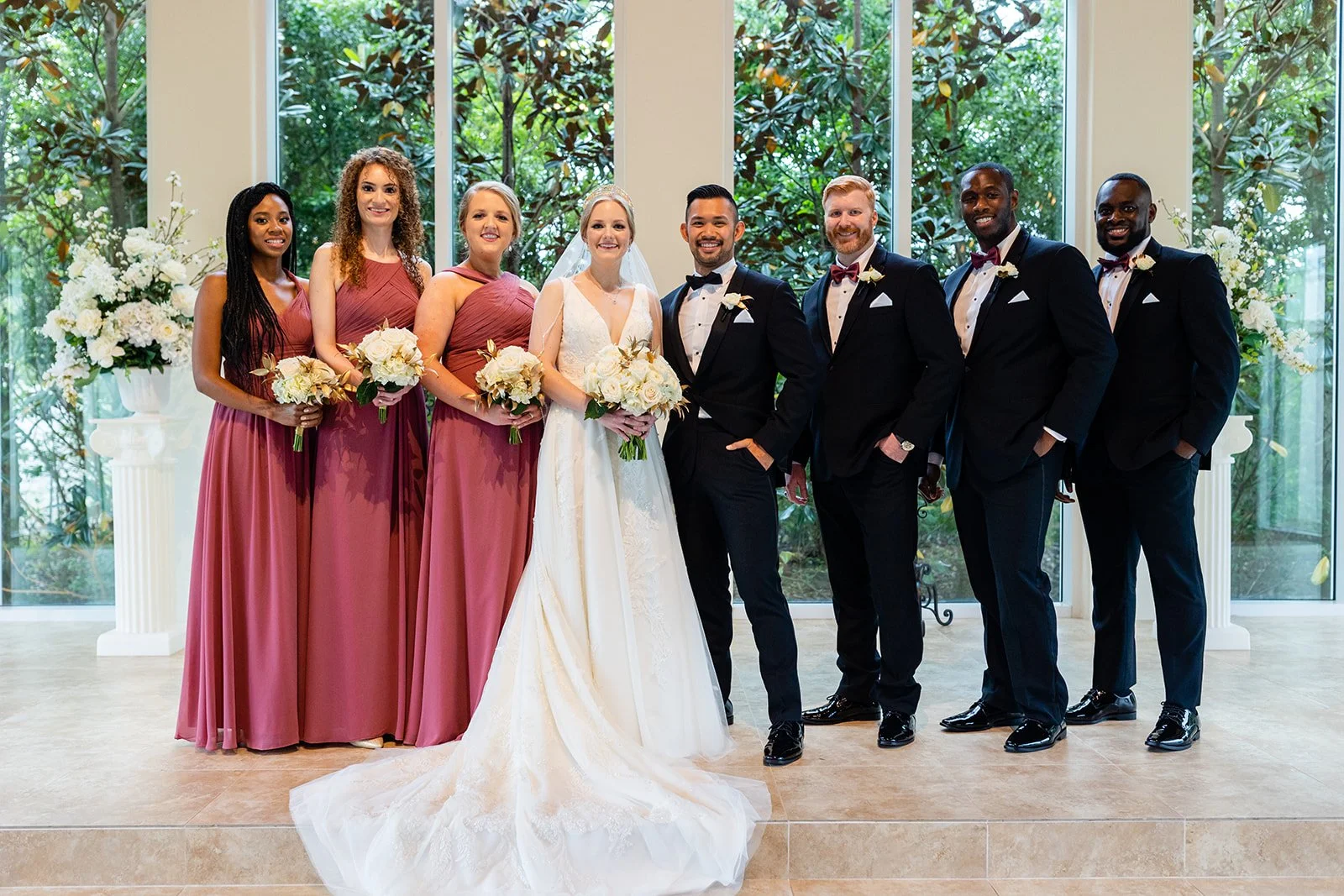 Bride, groom, and wedding party standing indoors with large glass windows showing greenery, all dressed in formal wedding attire, holding bouquets and smiling.