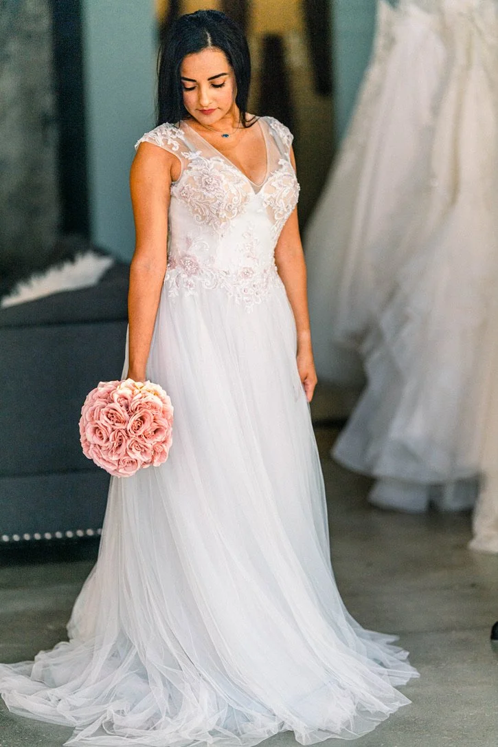 A bride in a white wedding dress holding a bouquet of pink roses, standing indoors.