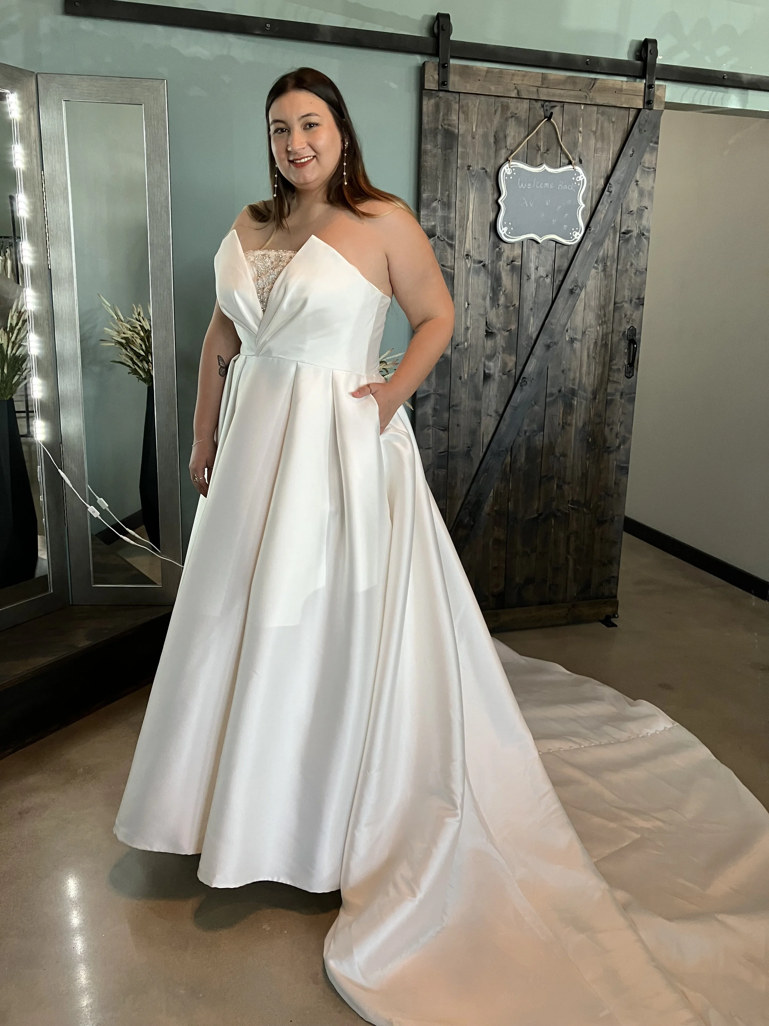 Woman in a white wedding gown with a strapless, detailed bust, standing indoors with a wooden sliding barn door behind her and a mirror with string lights to her left.