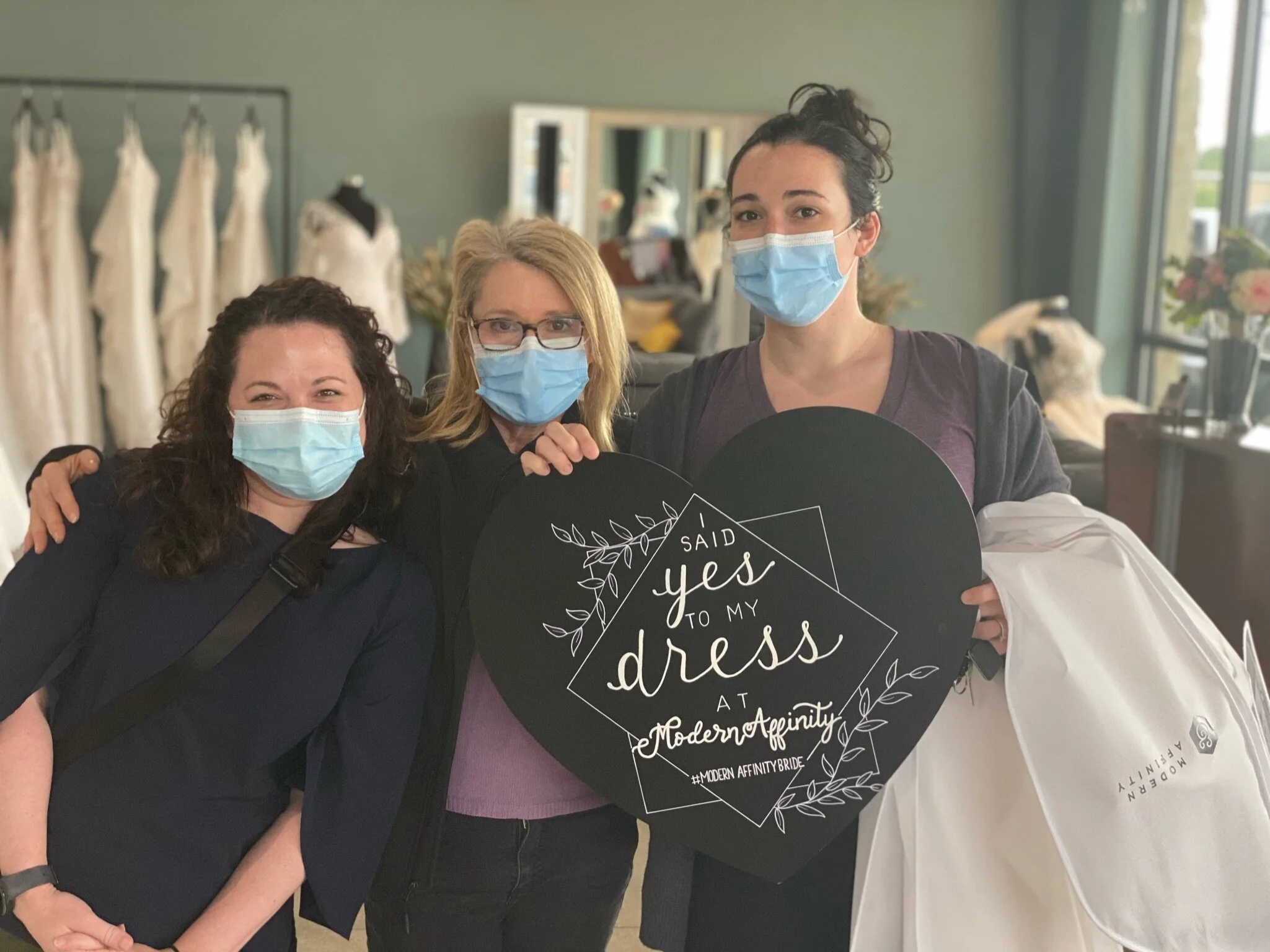 Three women wearing face masks standing inside a bridal shop, holding a heart-shaped sign that says 'Said yes to my dress at Modern Affinity'.
