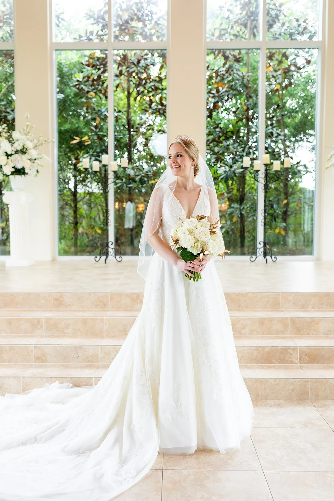 A bride in a white wedding dress holding a bouquet of white roses, standing in a bright indoor setting with large windows and greenery outside.