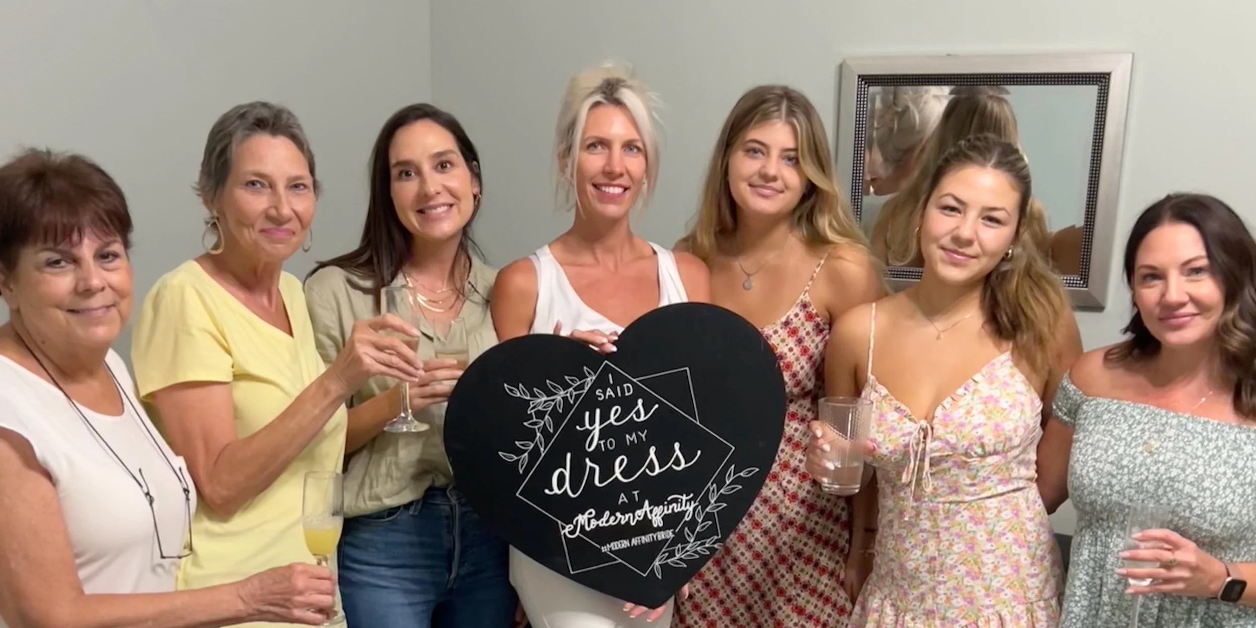 Group of women celebrating at a bridal shower, holding drinks and a black sign reading 'I said yes to my dress at Modern Affinity'.