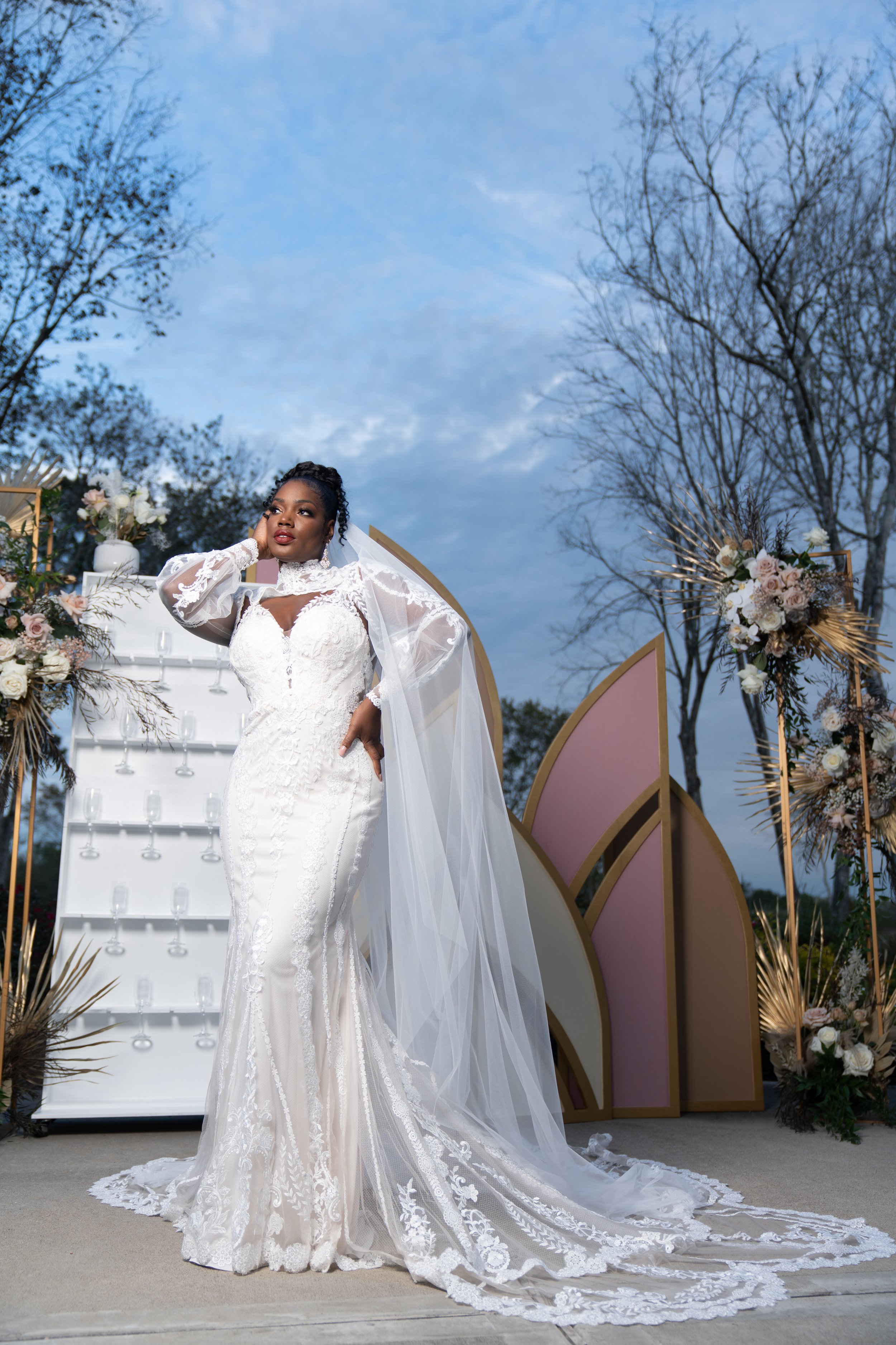 A bride in a white wedding gown with a long train, lace details, and a veil, standing outdoors during twilight with a decorated backdrop and floral arrangements.
