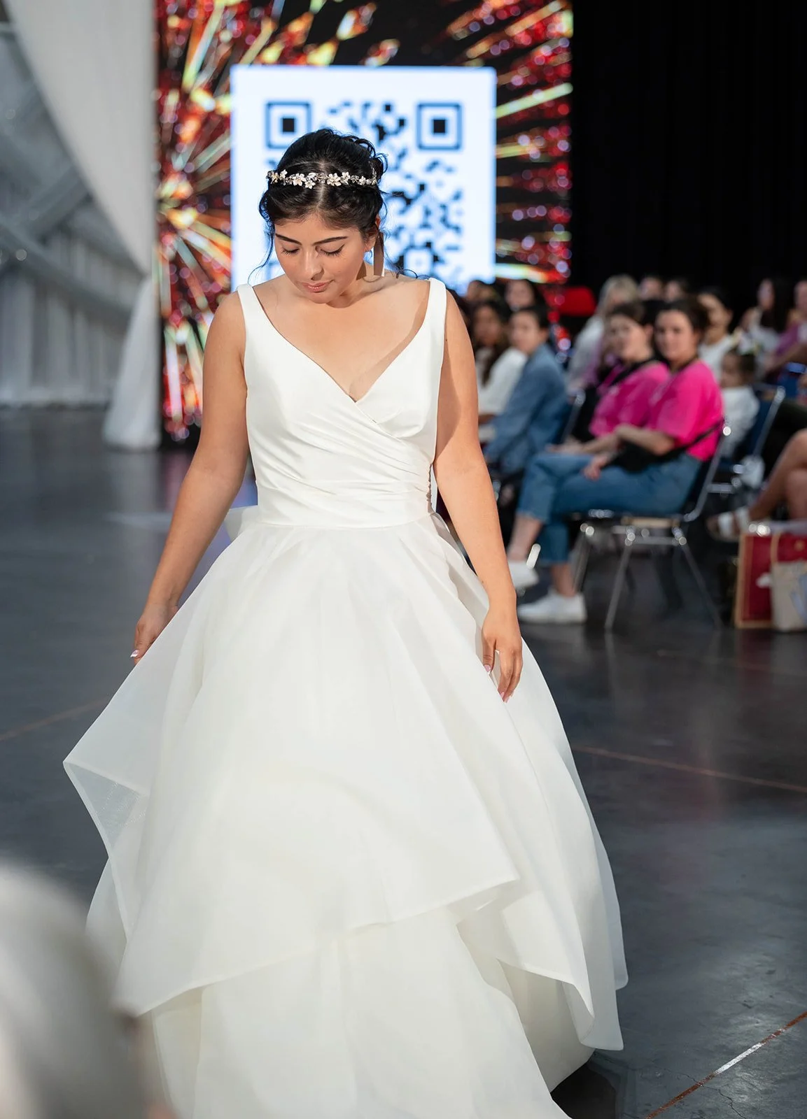 A woman in a white wedding dress walking on a stage with an audience in the background and a QR code on a screen behind her.