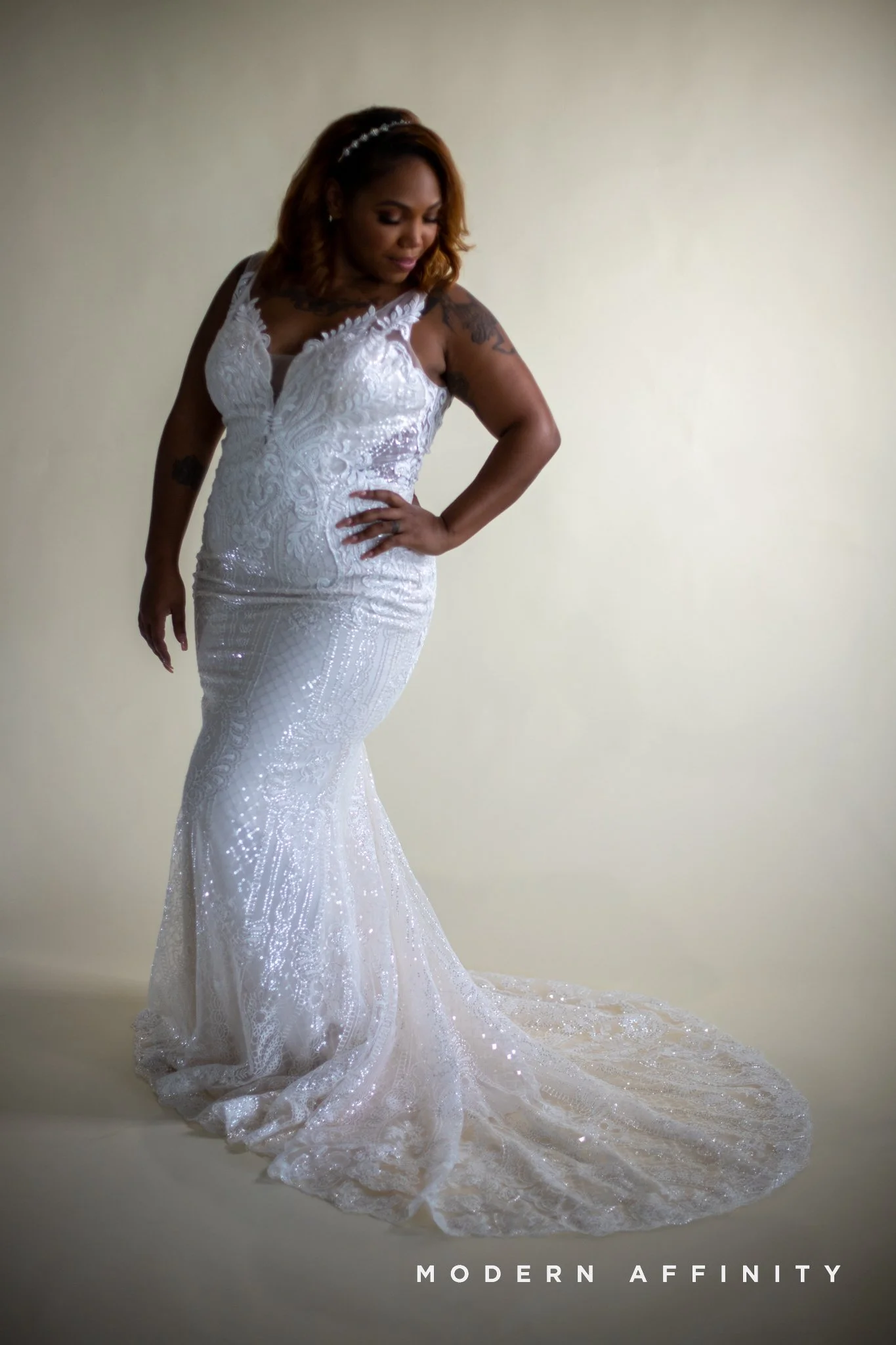 A woman in a white wedding gown with lace and embroidery details, standing against a plain background, looking down with her hand on her waist.