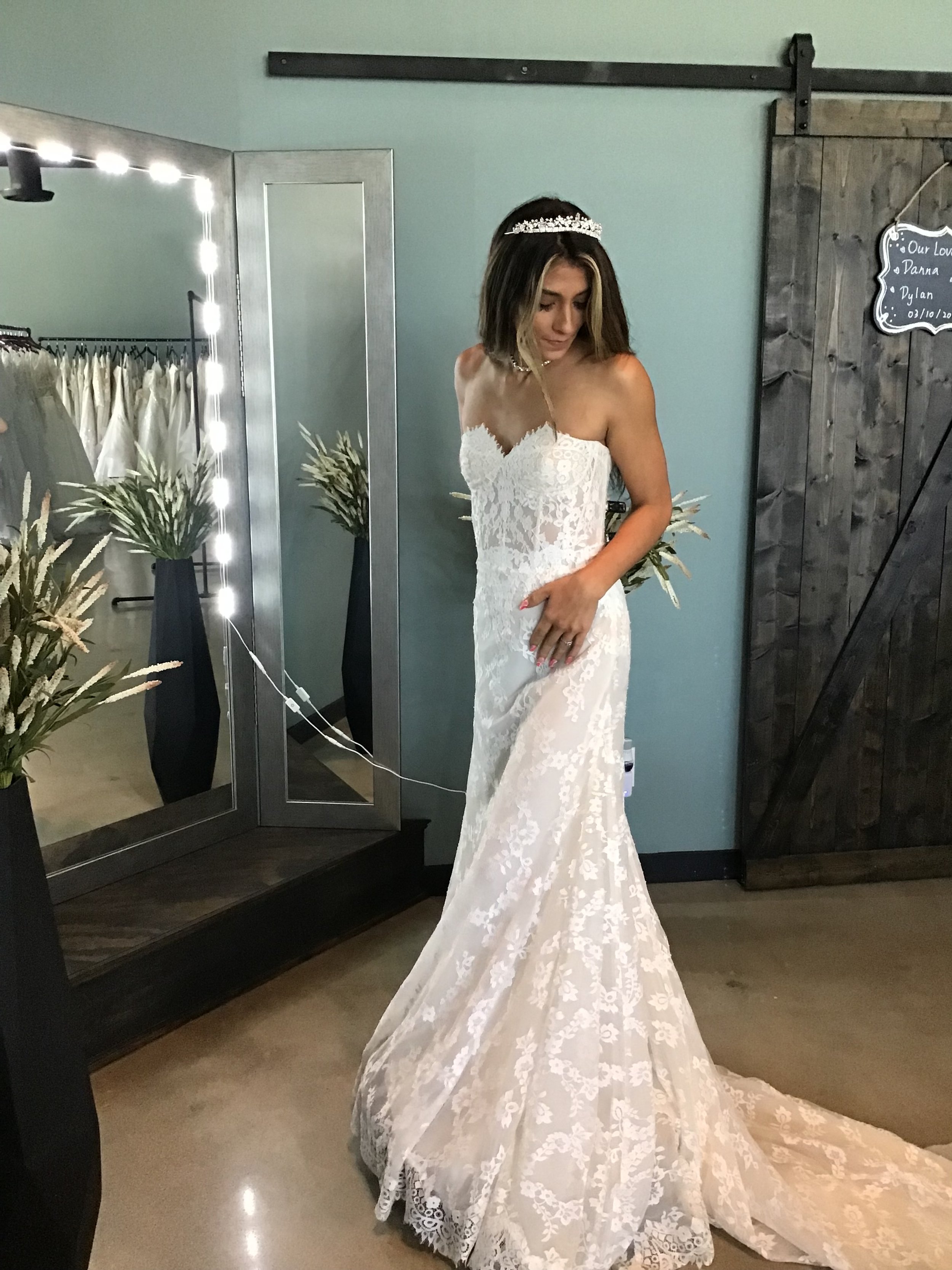 A woman in a white lace wedding dress with a strapless sweetheart neckline and a train, trying on the dress in a bridal shop fitting room.