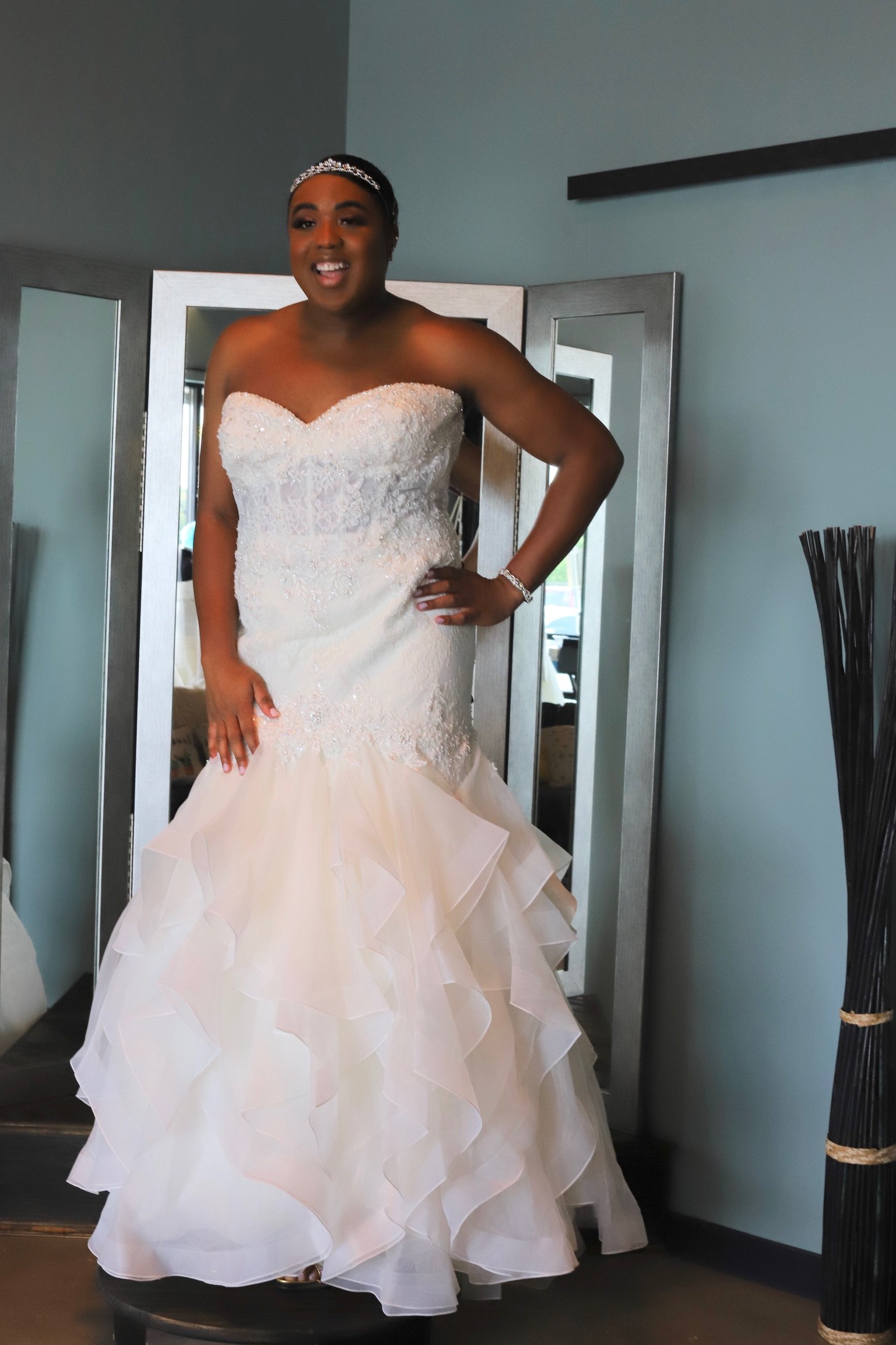A woman in a white wedding dress with a strapless, fitted bodice and a ruffled skirt stands on a stool in front of a mirror, smiling.