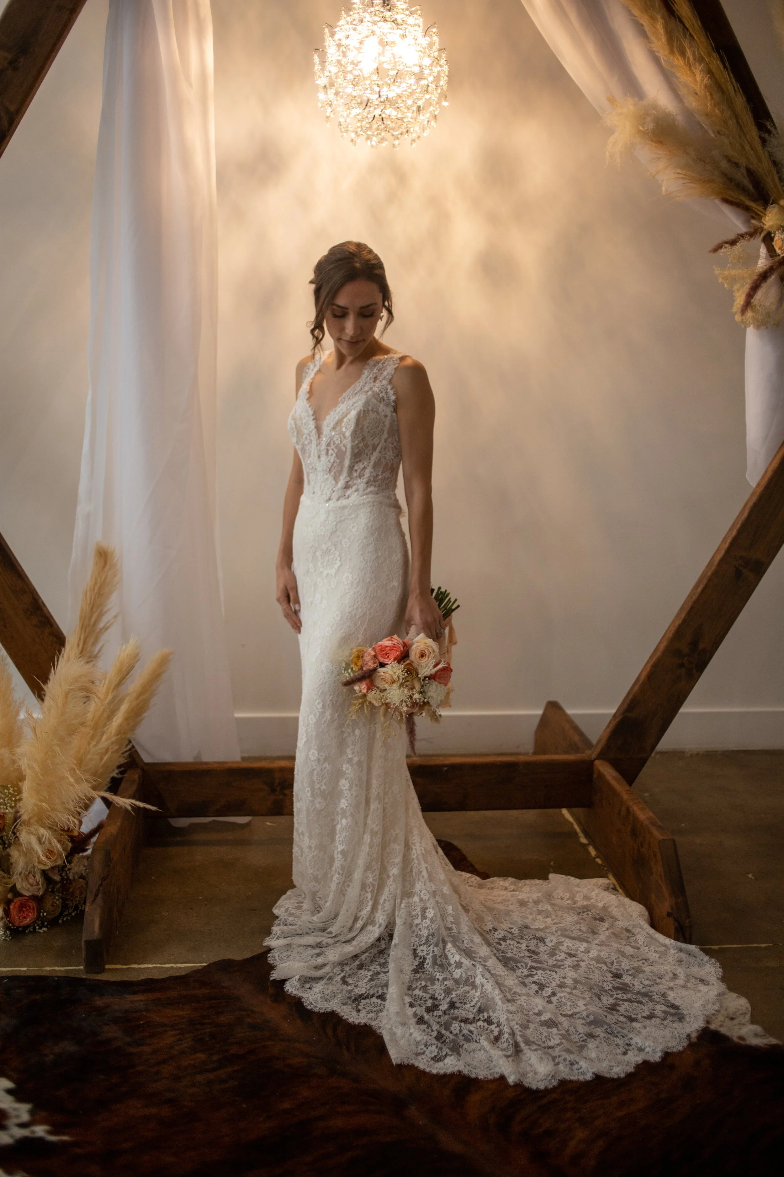 Bridal portrait of a bride in a lace wedding gown, holding a bouquet of pink and white roses, standing in a decorated setting with a chandelier overhead and floral arrangements.