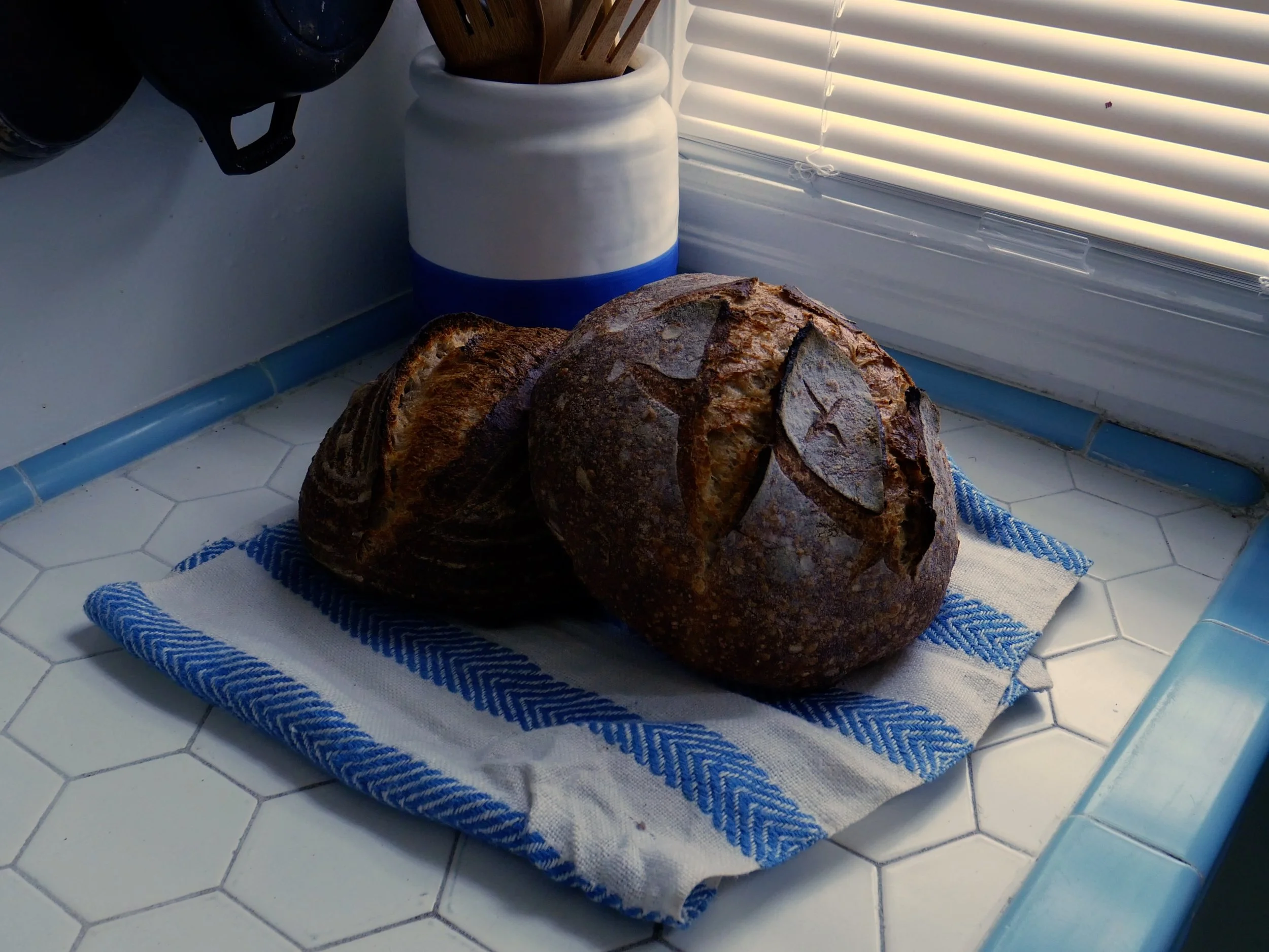 Two rustic loaves of bread on a striped blue and white towel on a white tiled countertop near a window with closed blinds, with kitchen utensils and a white ceramic container nearby.