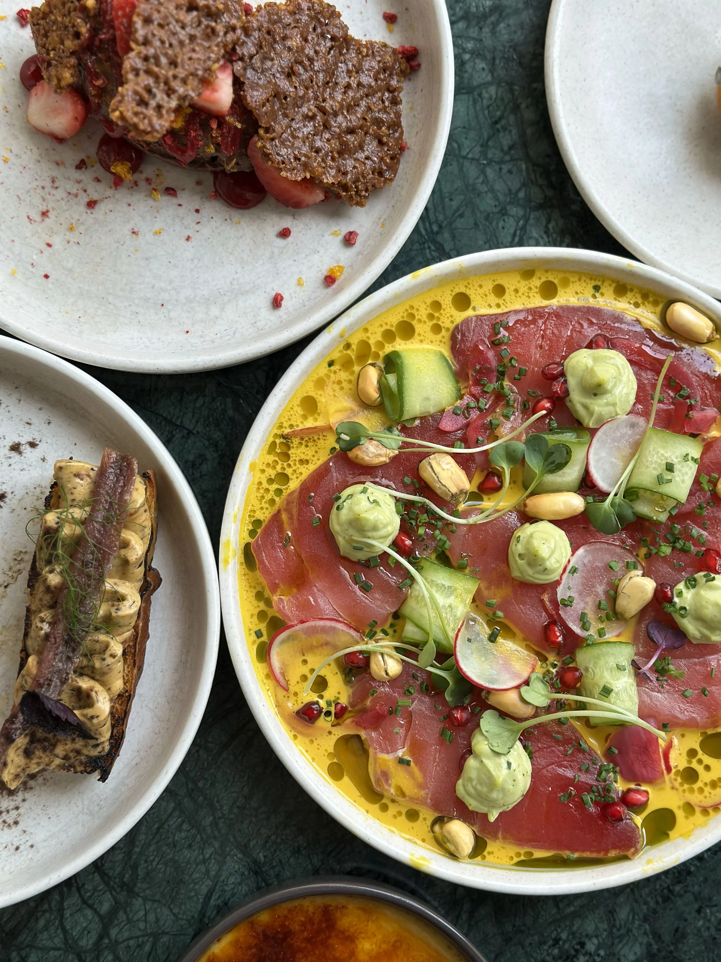 Close-up of a colorful plate of yellow-tinted dish with slices of raw fish, green sauce, radish slices, cucumber, chives, pea shoots, pomegranate seeds, with a yellow oil drizzle. 
