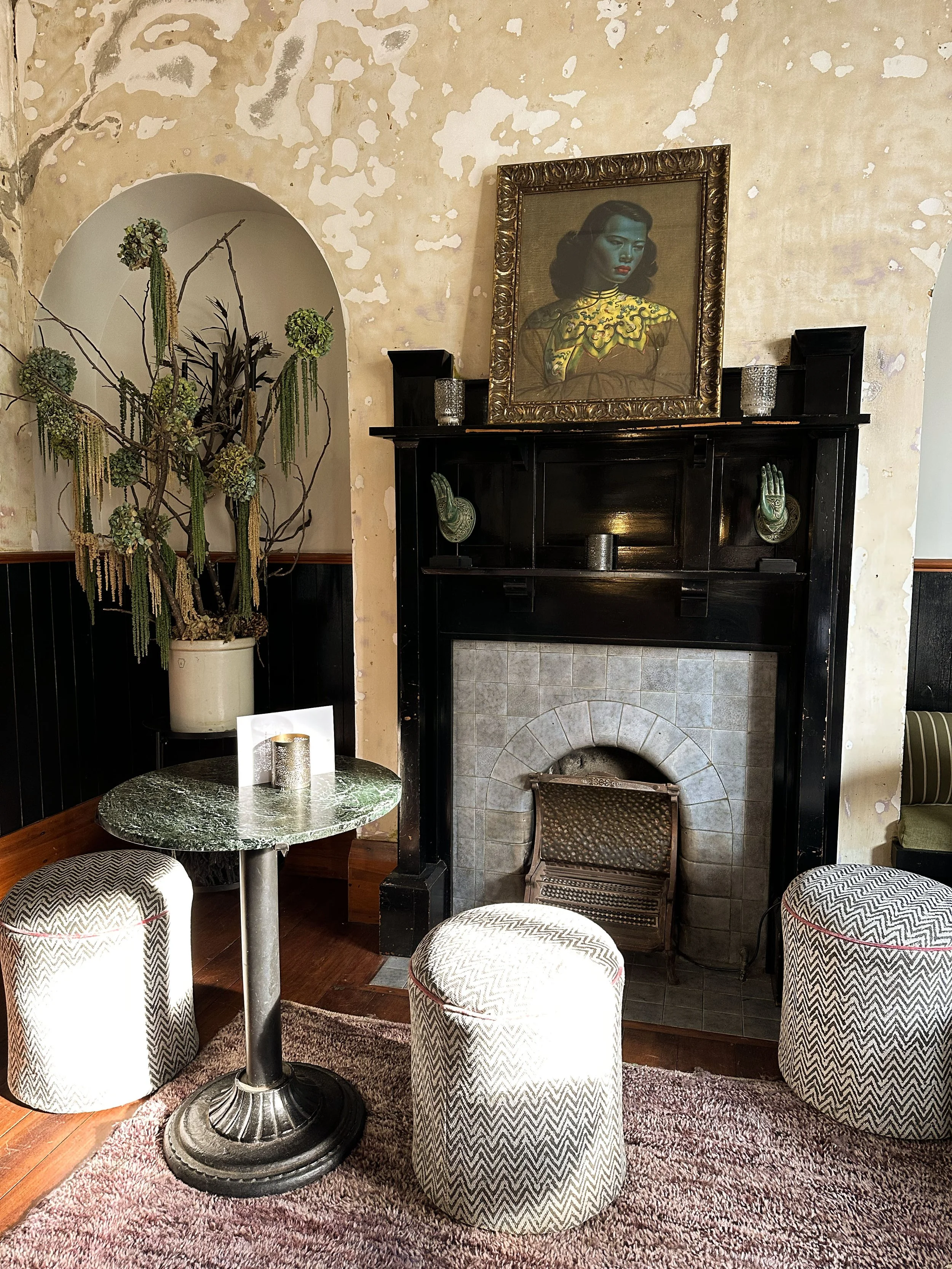 A cozy sitting area with a small round marble table, three patterned ottomans, a fireplace with black mantel, and a framed portrait of a woman above the mantel, with a decorative plant on the left side.