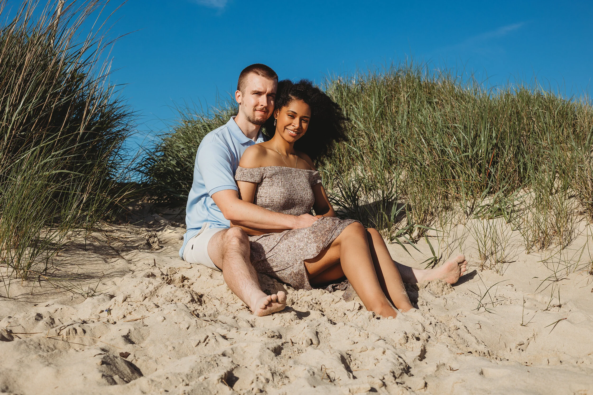 A couple sitting on sandy beach with green grass and blue sky in the background. The woman has curly black hair and is wearing an off-shoulder dress. The man has short hair and is wearing a light blue shirt and shorts.
