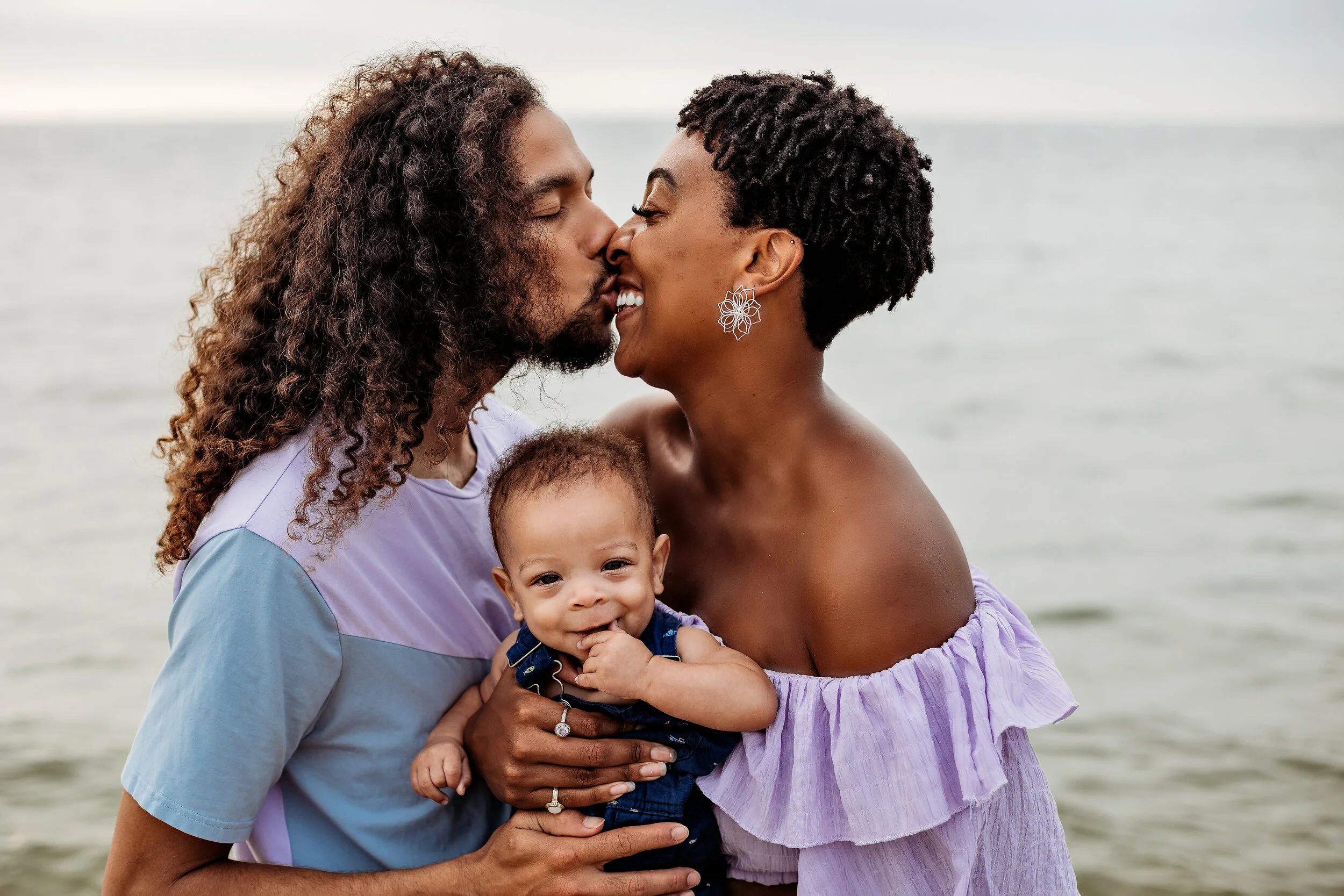 A couple is kissing while holding a young child at the beach with water in the background.
