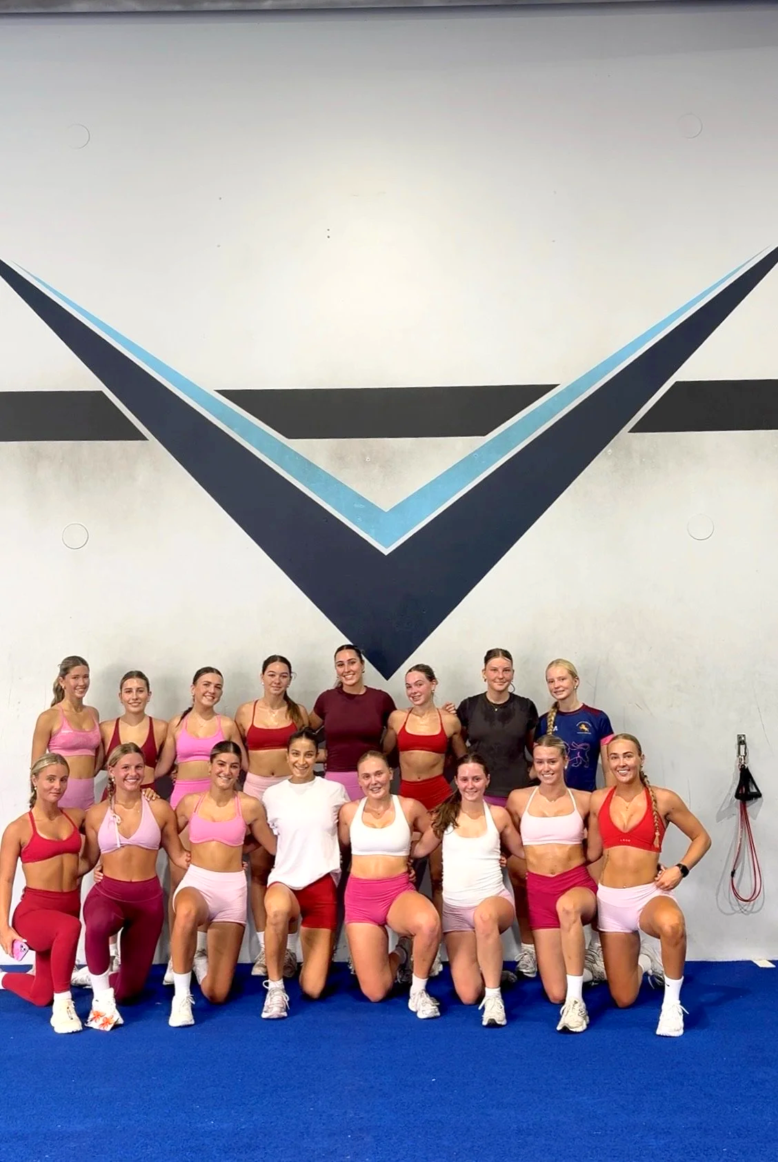 Group of 15 women in athletic sportswear standing and kneeling on a blue gym floor, posing for a photo in front of a wall with large geometric design. The women are smiling, some with arms around each other.