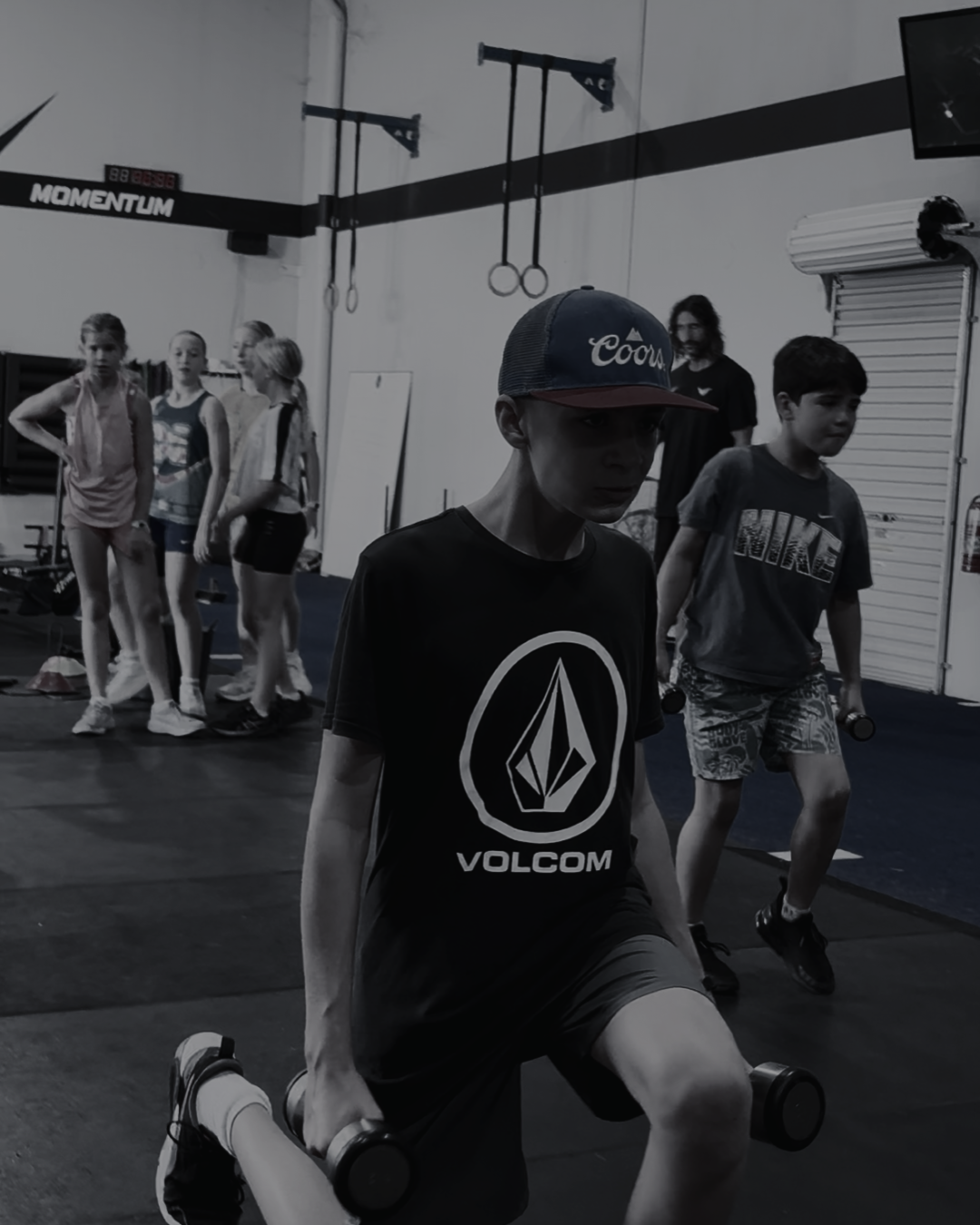 Young boy kneeling and holding dumbbells in a gym, with other children and a coach in the background.