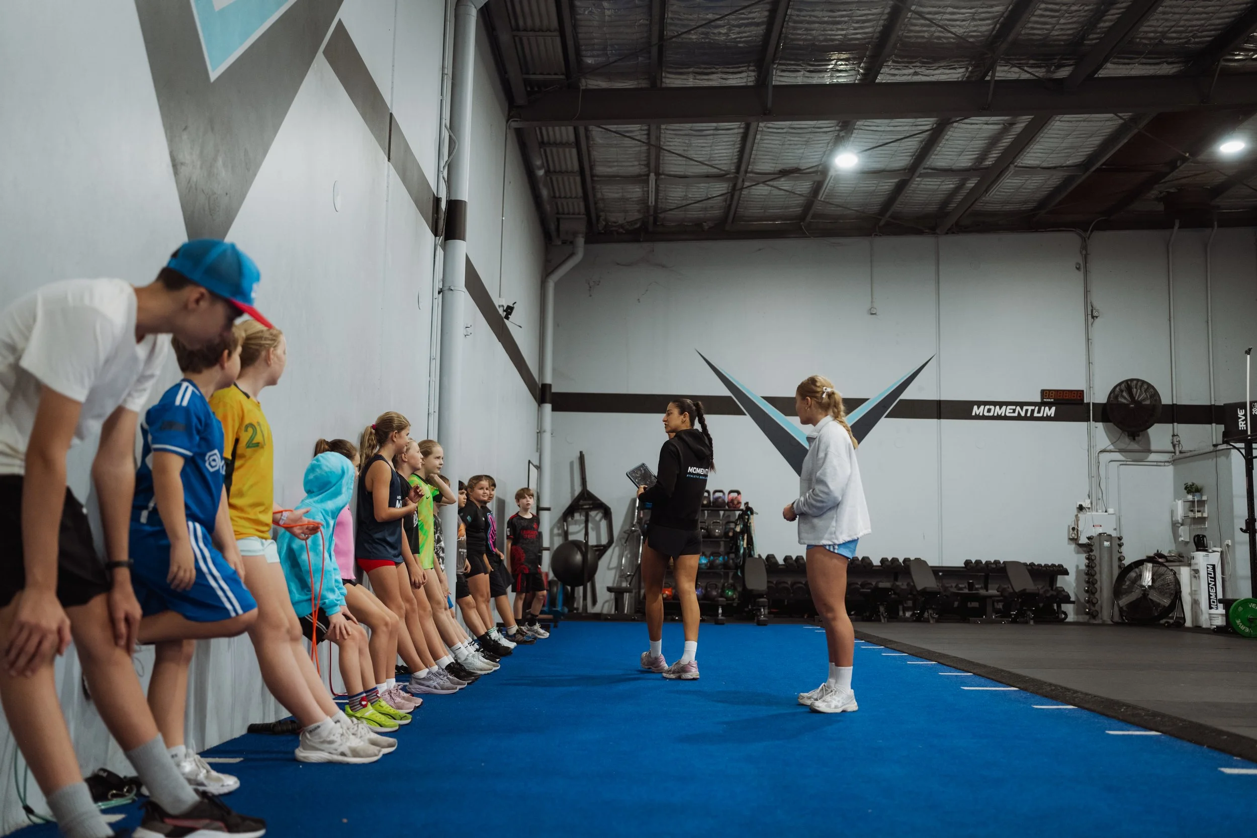 A group of children standing in a gym, listening to an instructor, with two women speaking to the class. The children are doing a stretching exercise and are dressed in athletic wear.