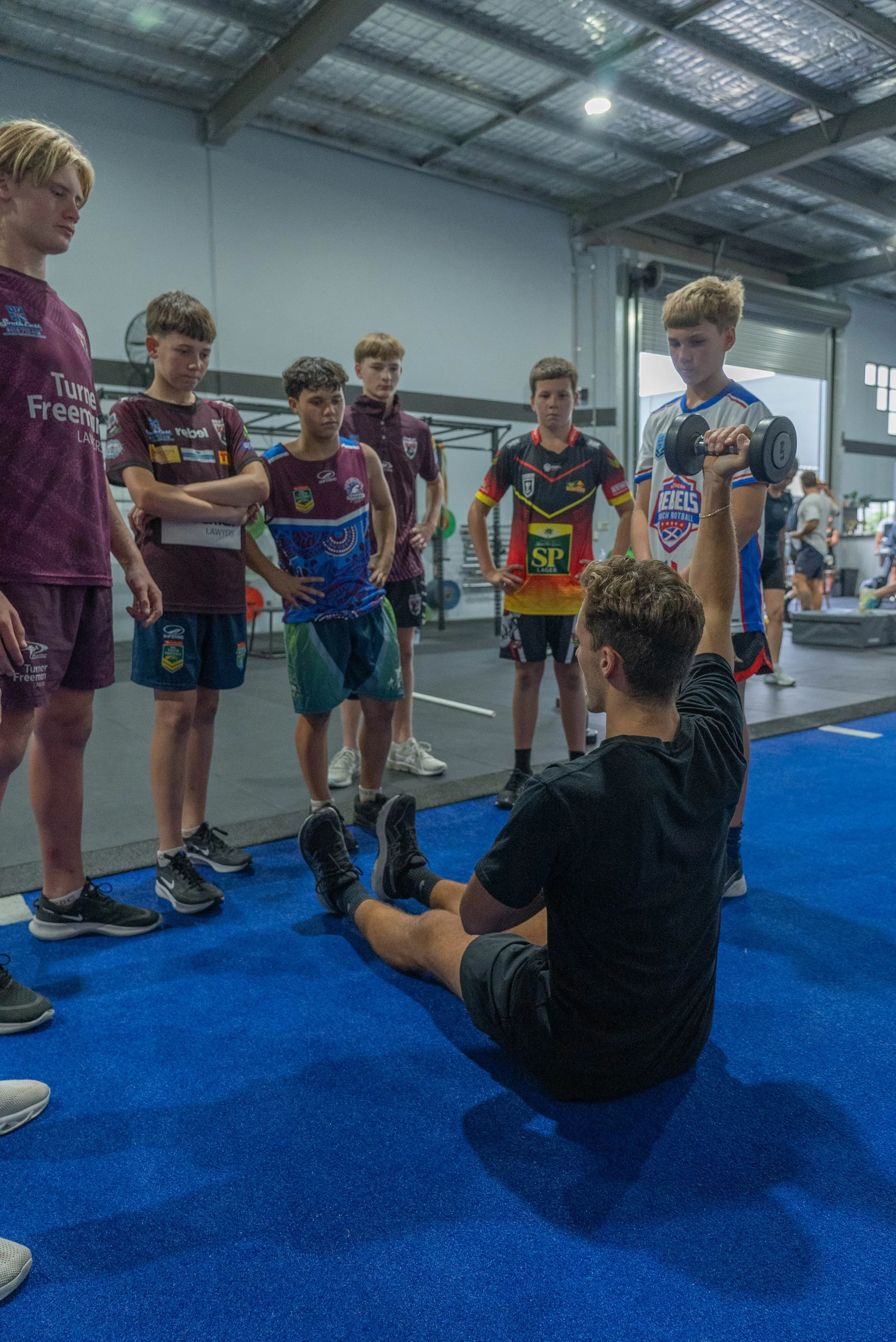 A group of young boys in sports uniforms standing around an adult male who is seated on the floor, holding a dumbbell. The scene takes place inside a gym with a blue mat on the floor and gym equipment in the background.