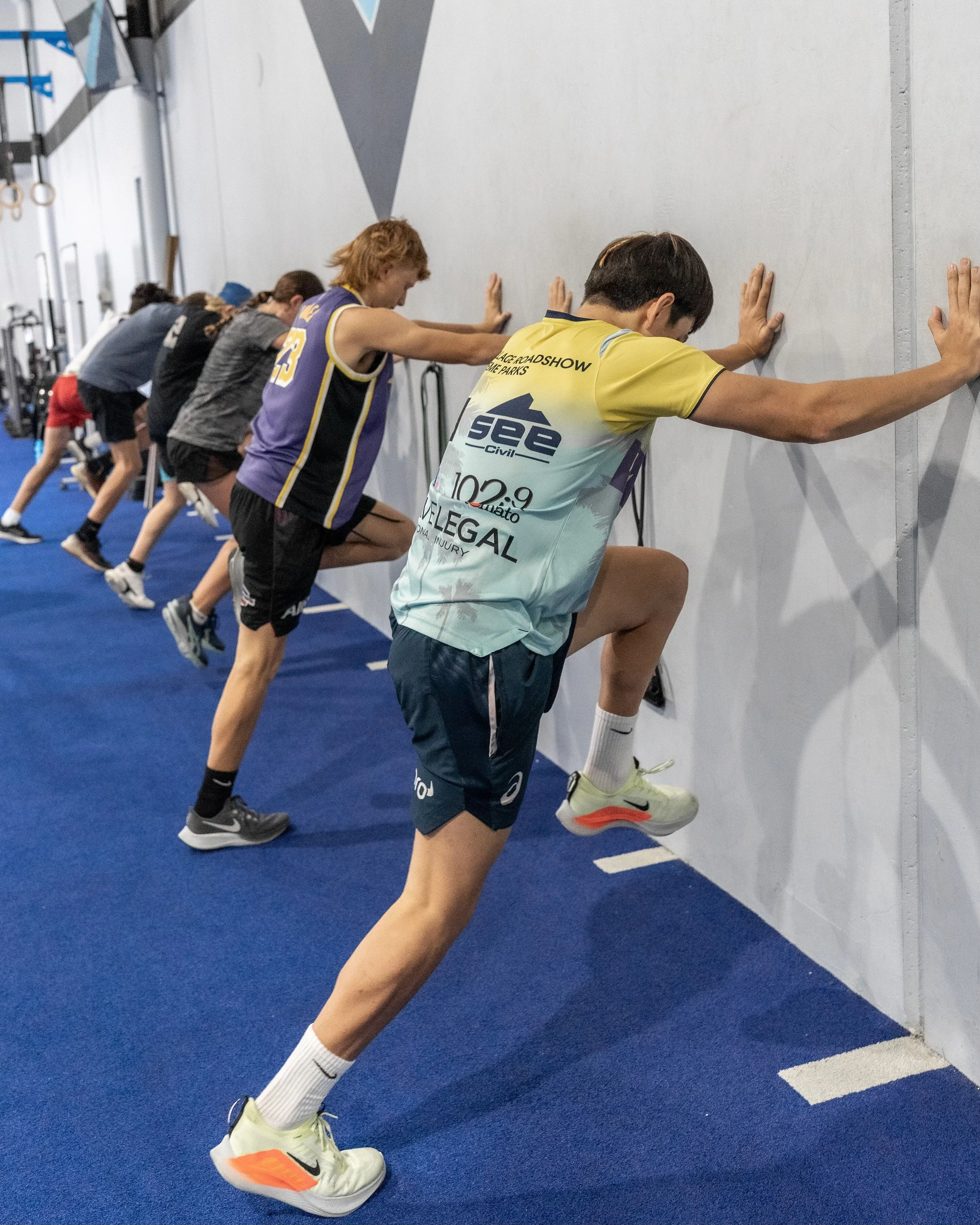 A group of people in athletic clothing doing wall push-ups in a gym with a blue carpeted floor and white walls. The focus is on the person in the foreground wearing a light-colored shirt, black shorts, and white and orange running shoes, with arms ex