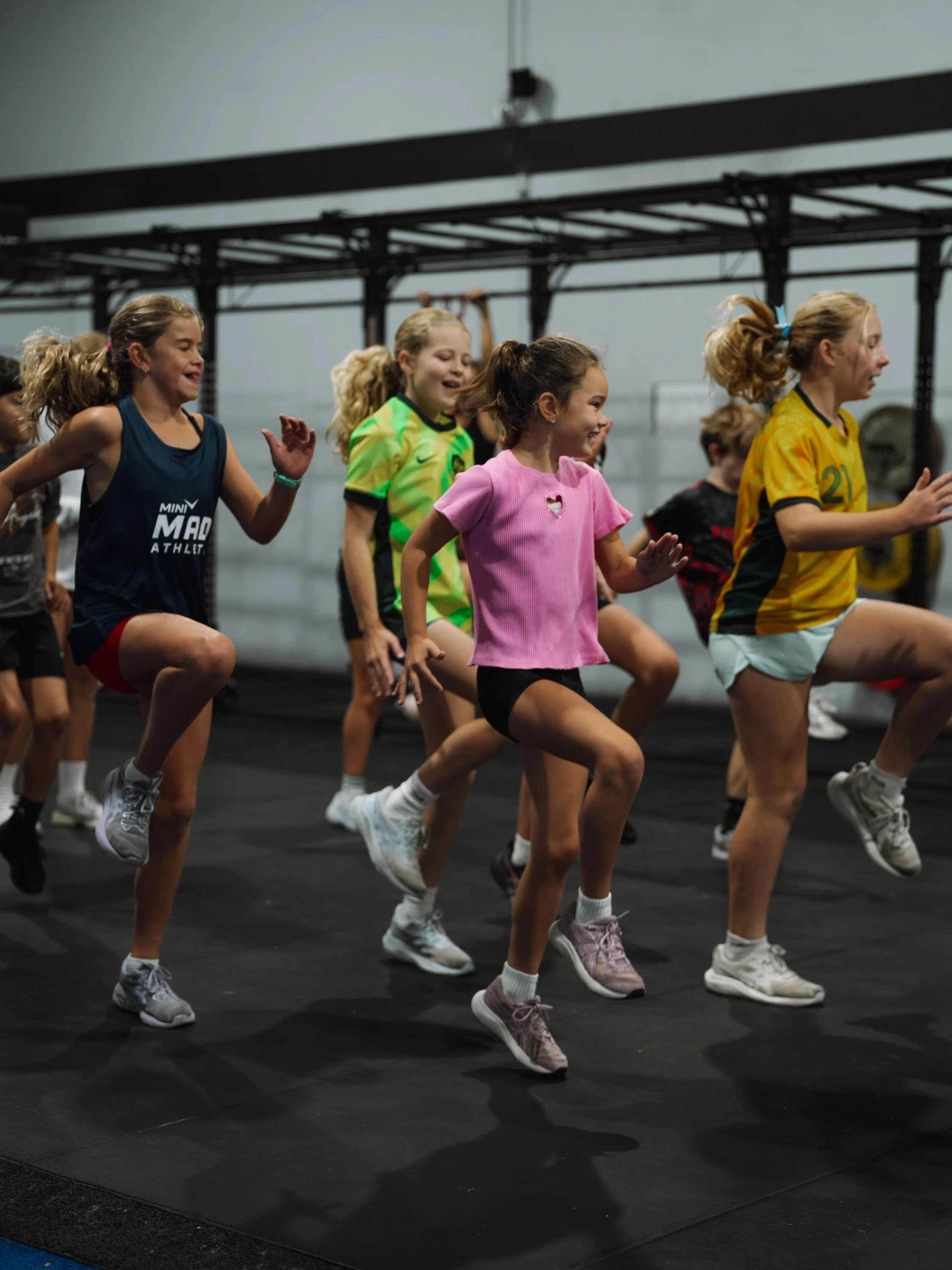 A woman with long red hair doing a wall squat exercise in a gym. She is wearing a pink sports bra, matching shorts, pink socks, and white athletic shoes, and is holding a dumbbell in each hand.