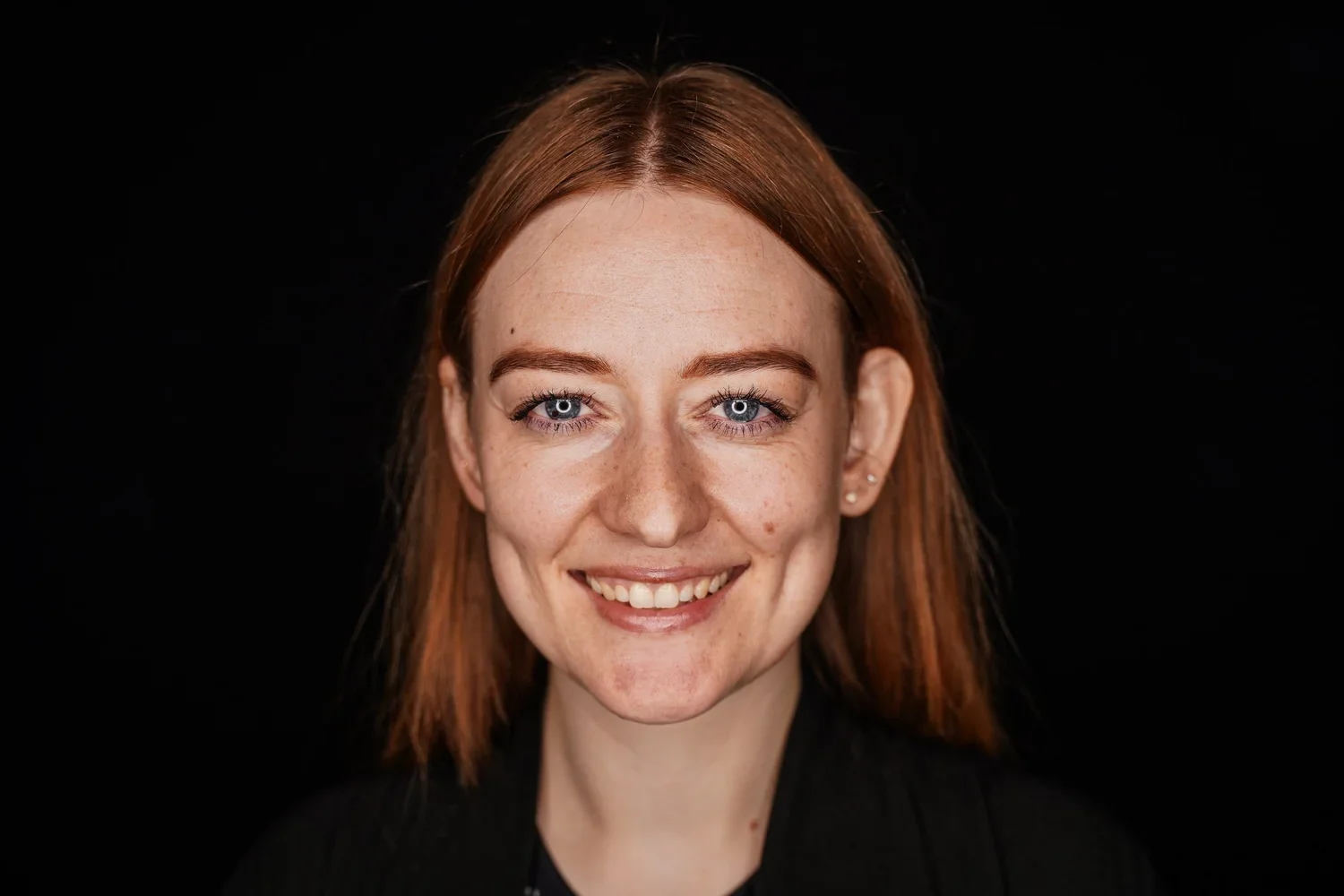 A young woman with red hair and blue eyes smiling against a black background.