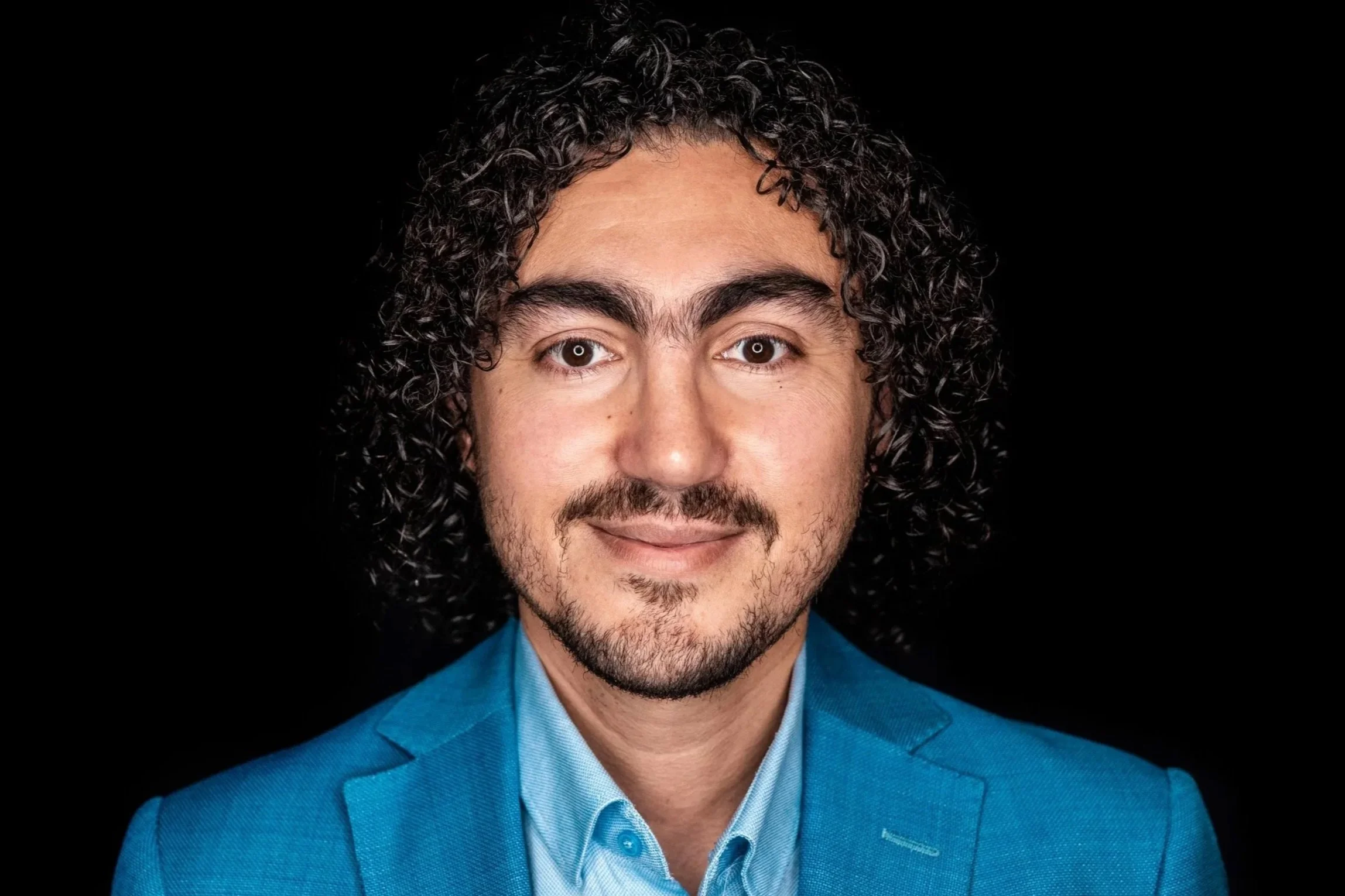 Close-up of a man with curly dark hair, beard, and mustache, wearing a blue suit and shirt, smiling at the camera against a black background.