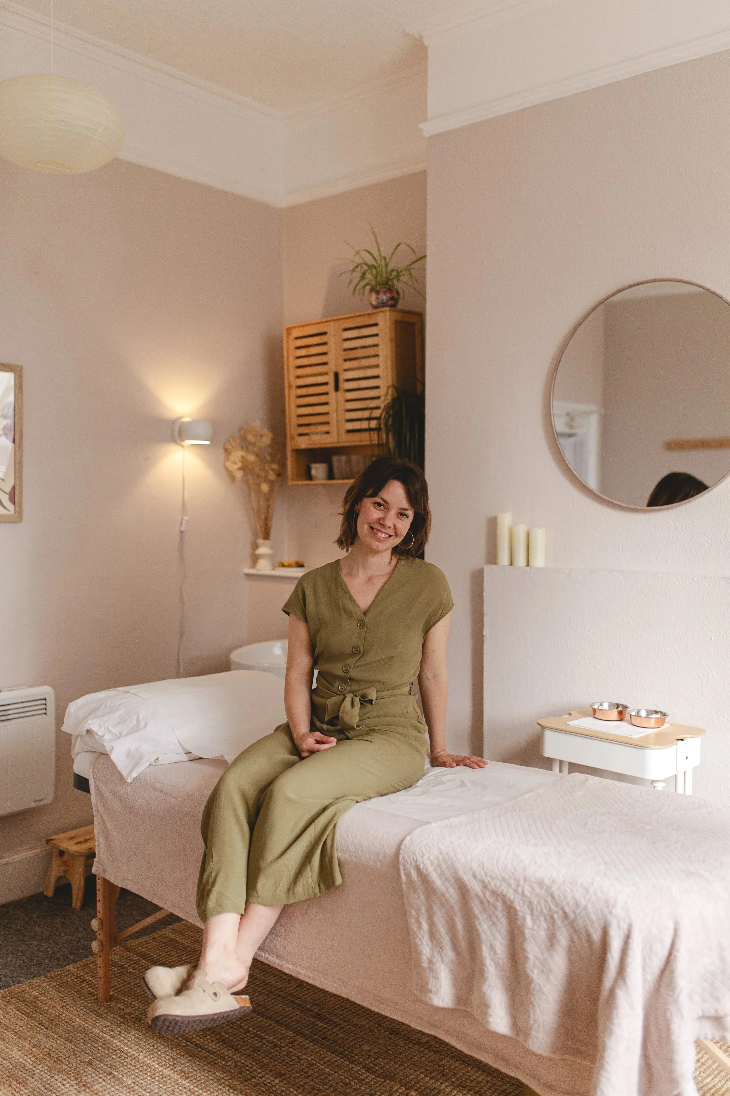 A young woman with short brown hair wearing a green jumpsuit and beige slippers, sitting on the edge of a bed in a softly lit bedroom, smiling at the camera. The room has a round mirror on the wall, a small white nightstand with candles, wooden shelves with plants, and a cozy, neutral color scheme.