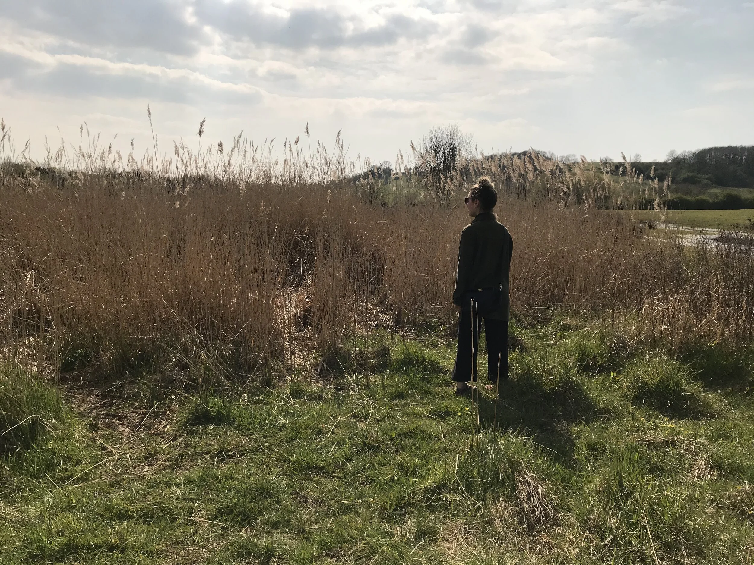 Person standing outdoors in a field with tall grass and reeds, wearing dark clothing and sunglasses, looking away from the camera during a partly cloudy day.
