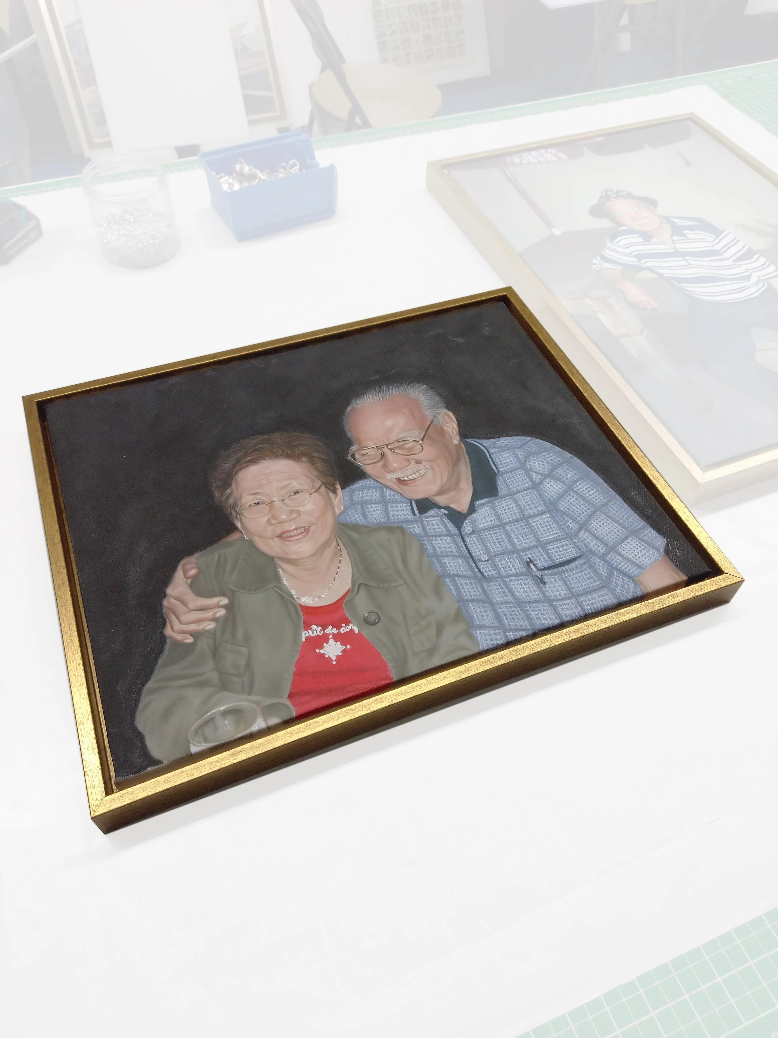 A framed photo of an elderly couple smiling and posing together, with the man placing his hand on the woman's shoulder, on a white table with craft supplies.