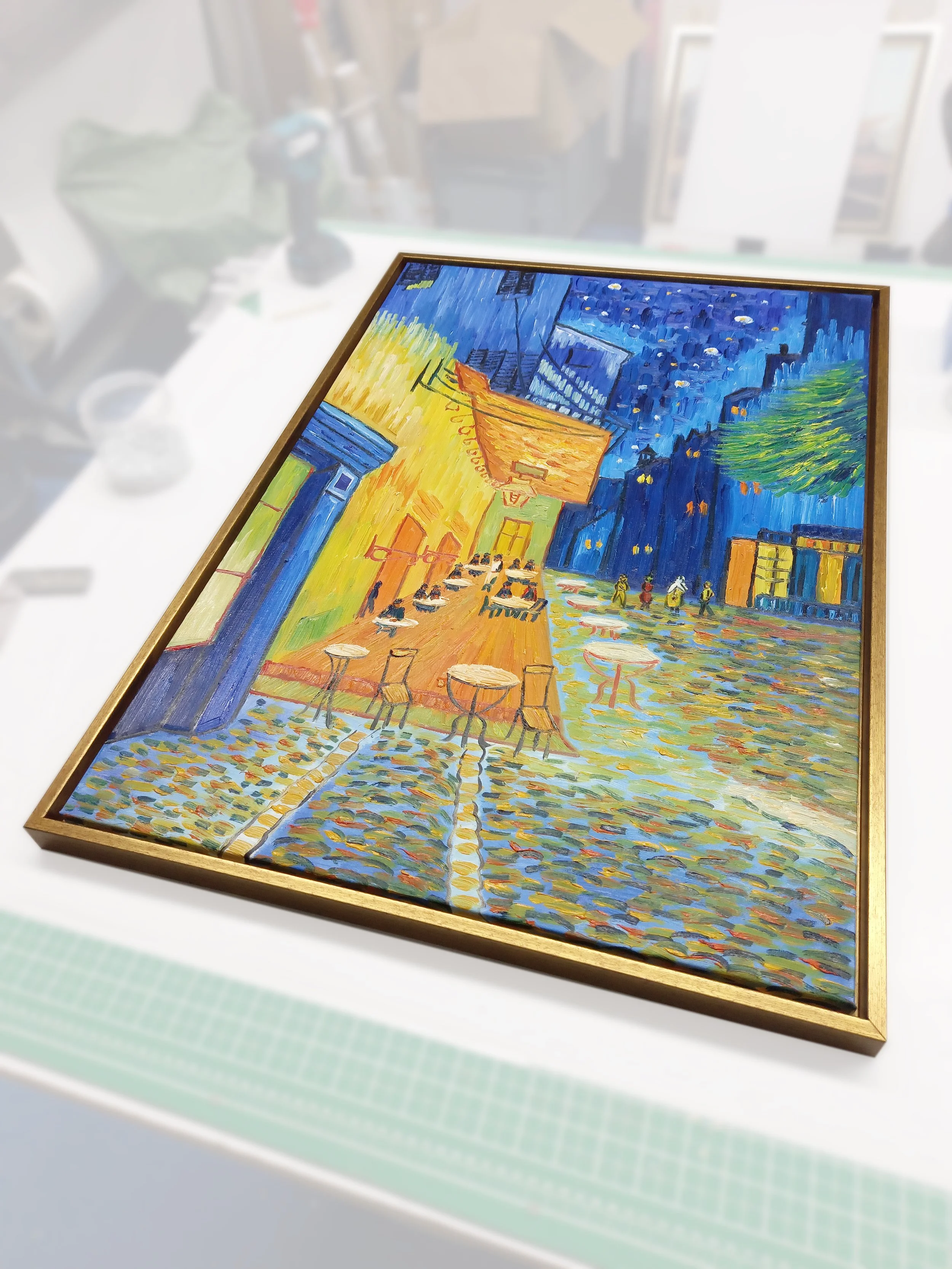 A framed painting of a city street scene at night, featuring a yellow building, outdoor cafe tables, people walking, and a starry sky with a crescent moon.