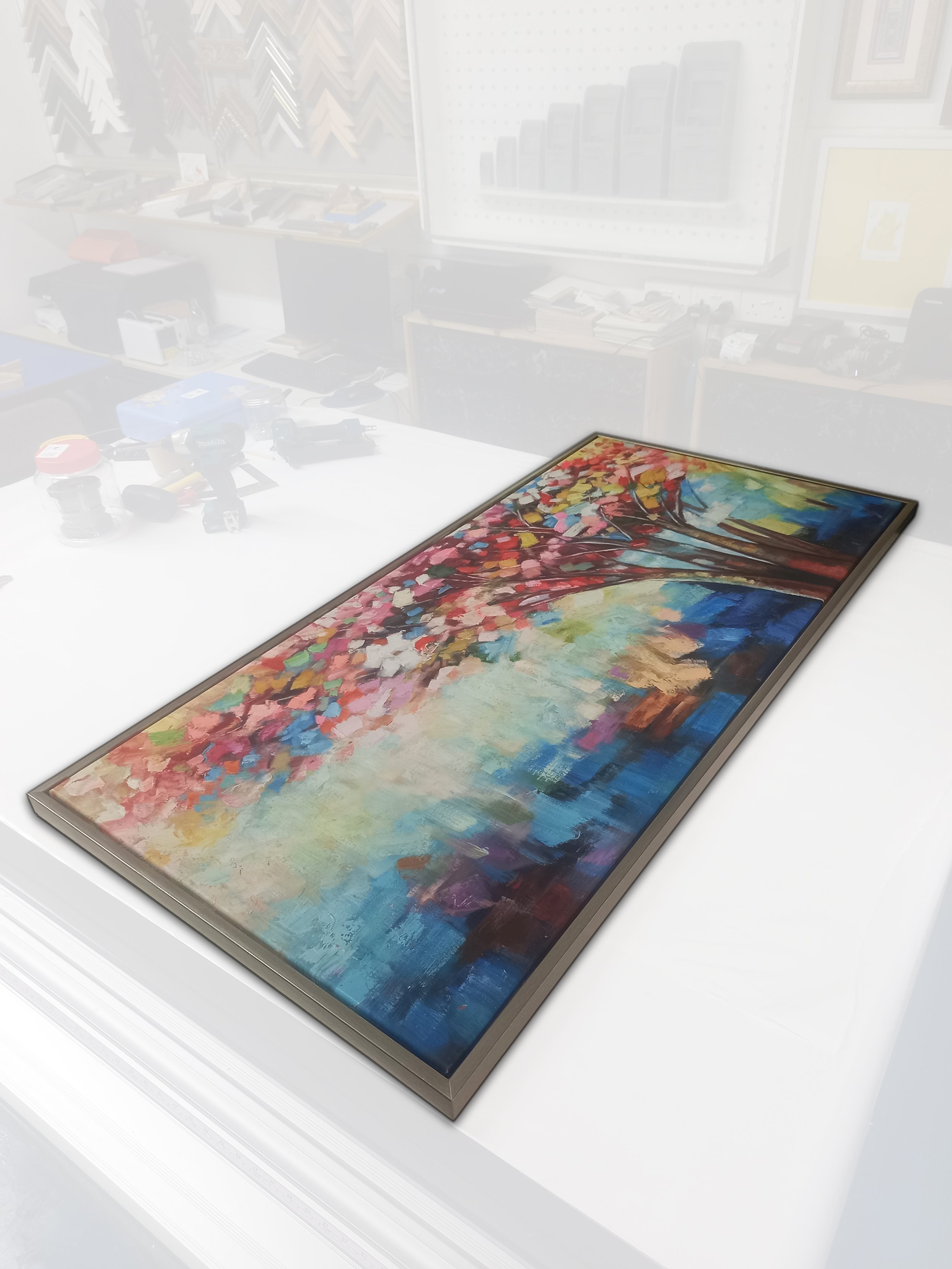 Colorful abstract painting on a rectangular canvas displayed on a white table in a gallery. The artwork features vibrant strokes and a tree with pink leaves on the left side, contrasted by dark and bright blue and white areas.
