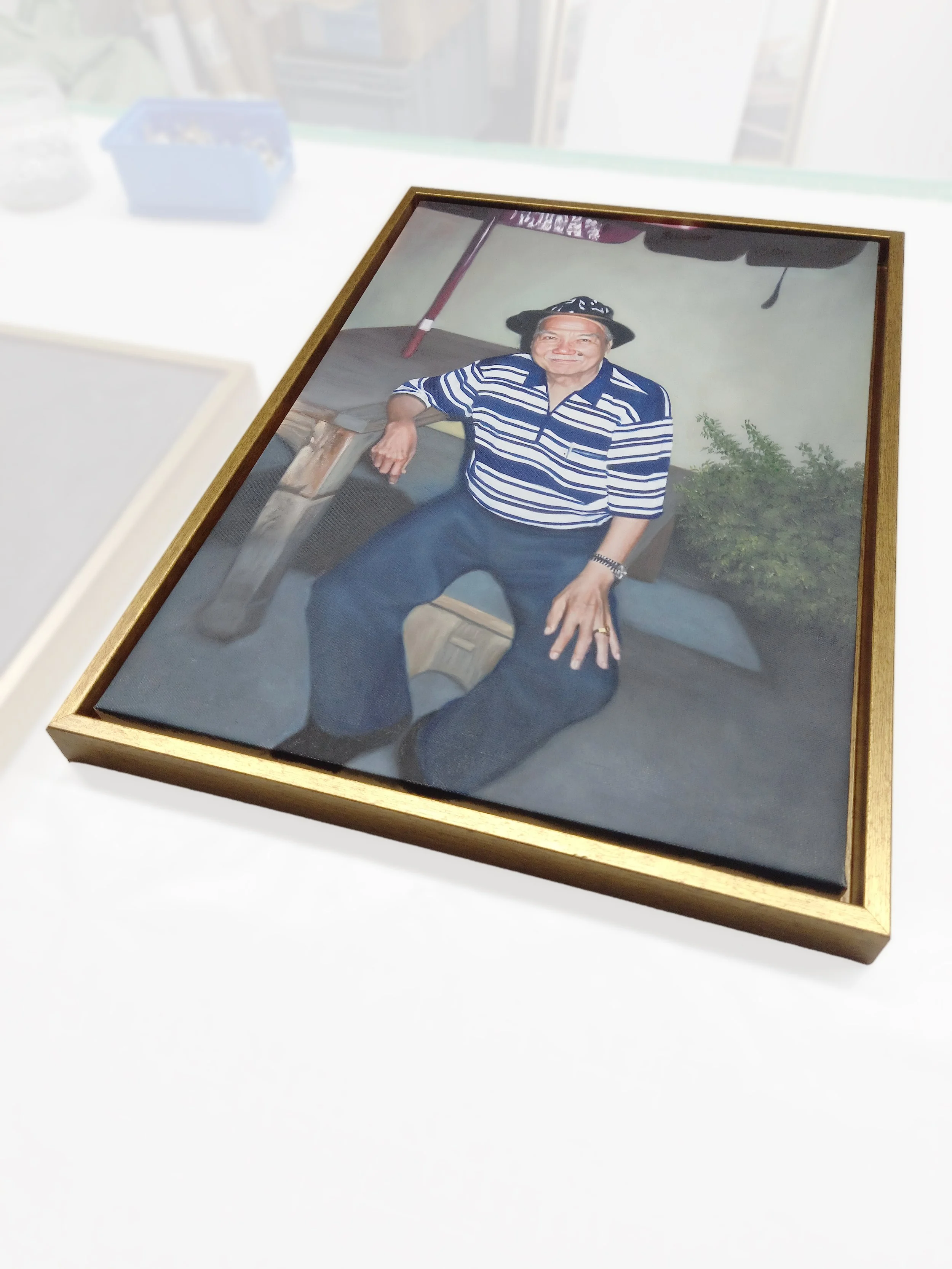 Framed photograph of an elderly man wearing a wide-brimmed hat and striped shirt, sitting outdoors with a table and plant in the background.