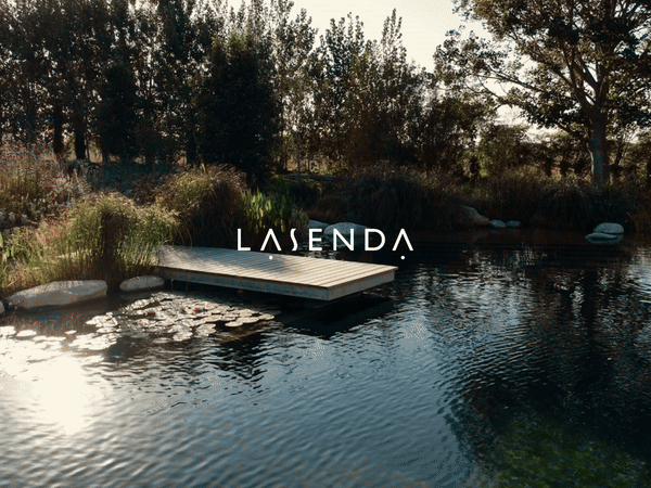 A small dock extends into a peaceful pond with trees and bushes along the shoreline, and the word 'LASENDA' displayed across the image.
