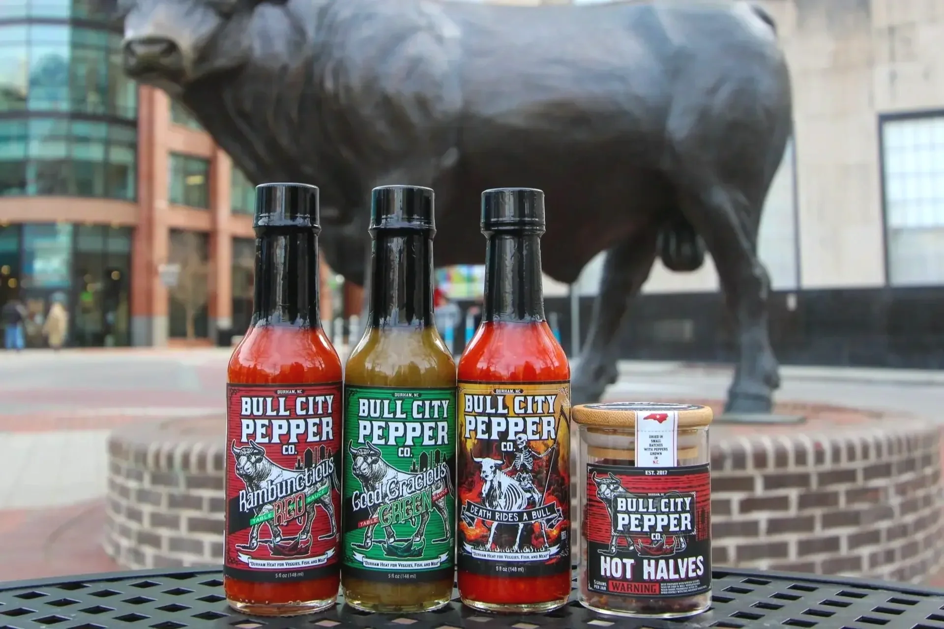 Four bottles and one jar of Bull City Pepper hot sauces and seasonings in front of a large bull statue outside