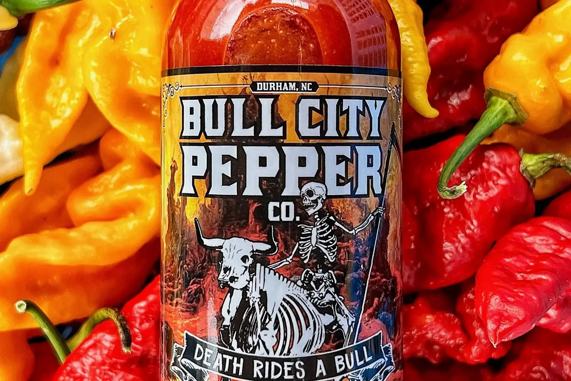 A bottle of Bull City Pepper Co. hot sauce surrounded by colorful bell and chili peppers, with a label featuring a skeleton riding a bull and the slogan "Death Rides a Bull".