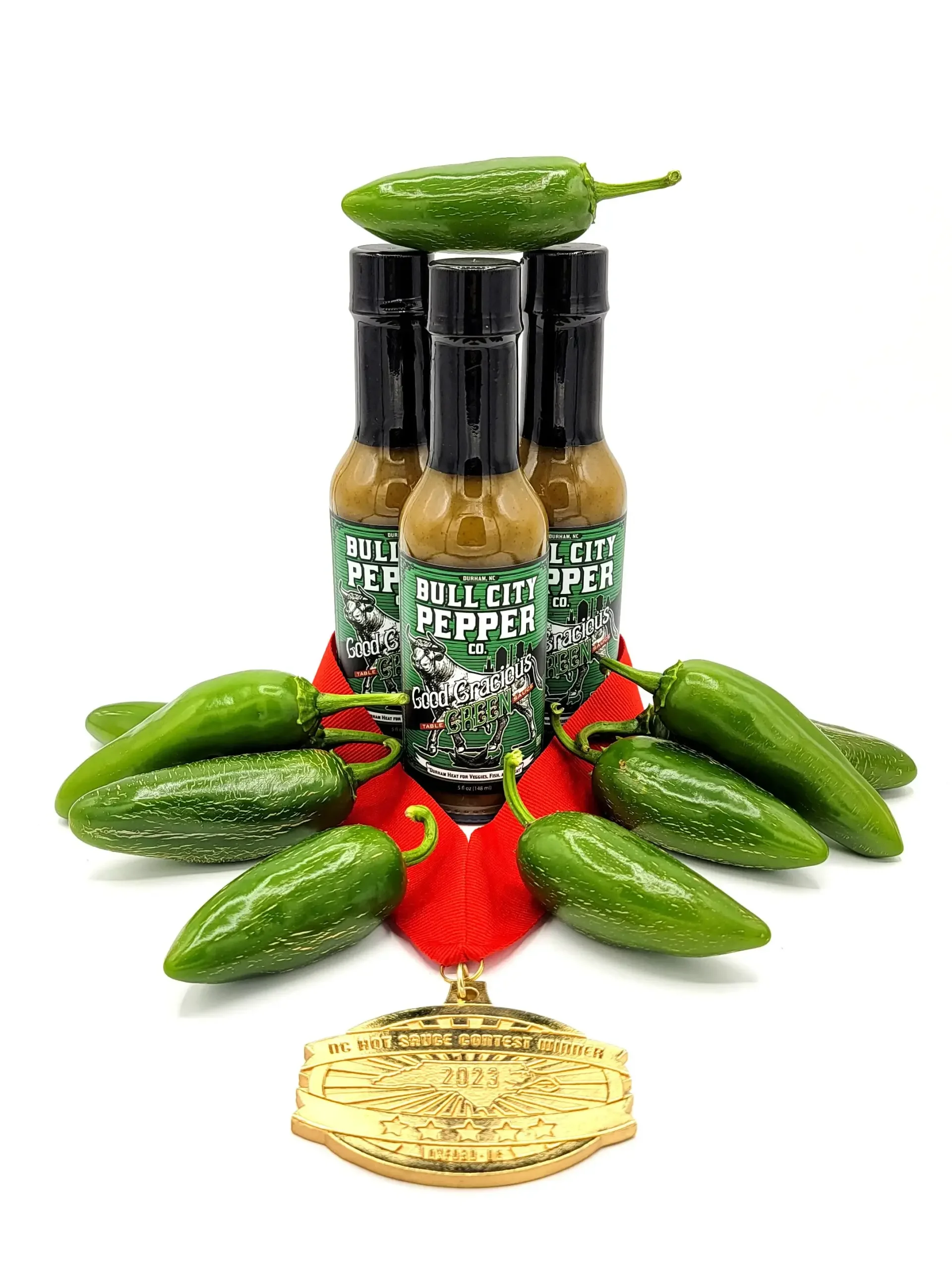 Stack of three bottles of Good Gracious Green Hot Sauce by Bull City Pepper Co. hot sauce surrounded by fresh jalapeño peppers, with a gold medal hanging from a red ribbon in front of the peppers.