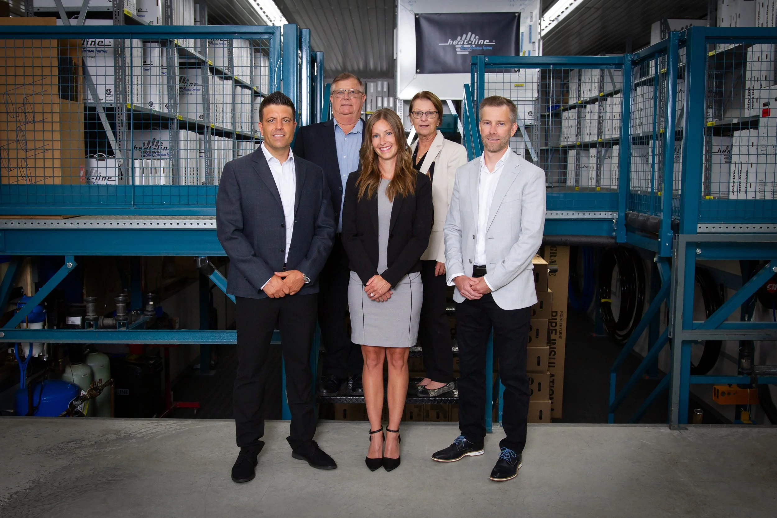 Group of six business professionals standing in an industrial or warehouse setting with metal shelving and equipment in the background.