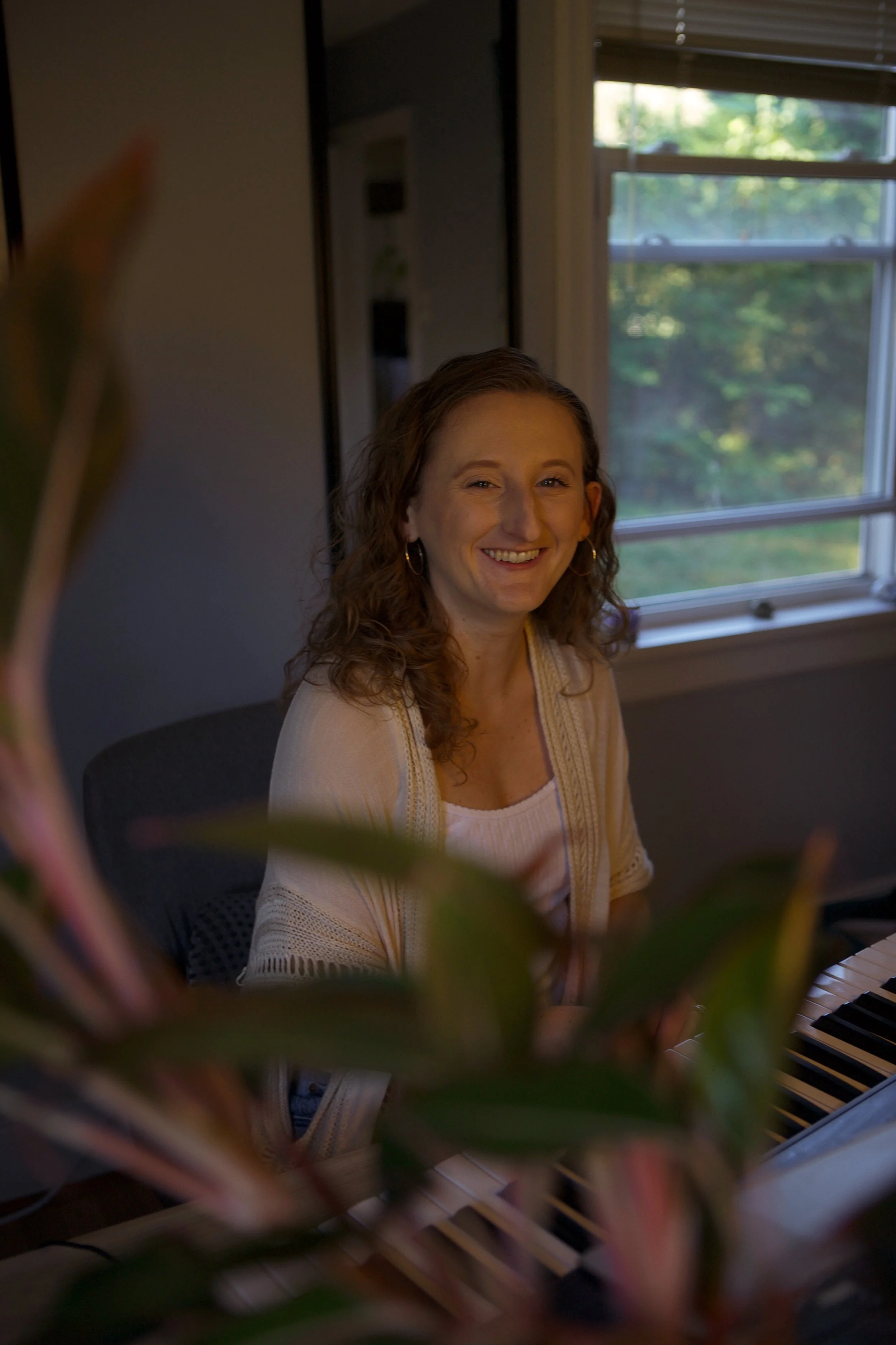 Woman with brown curly hair smiling while sitting at a piano with a window showing green trees in the background.