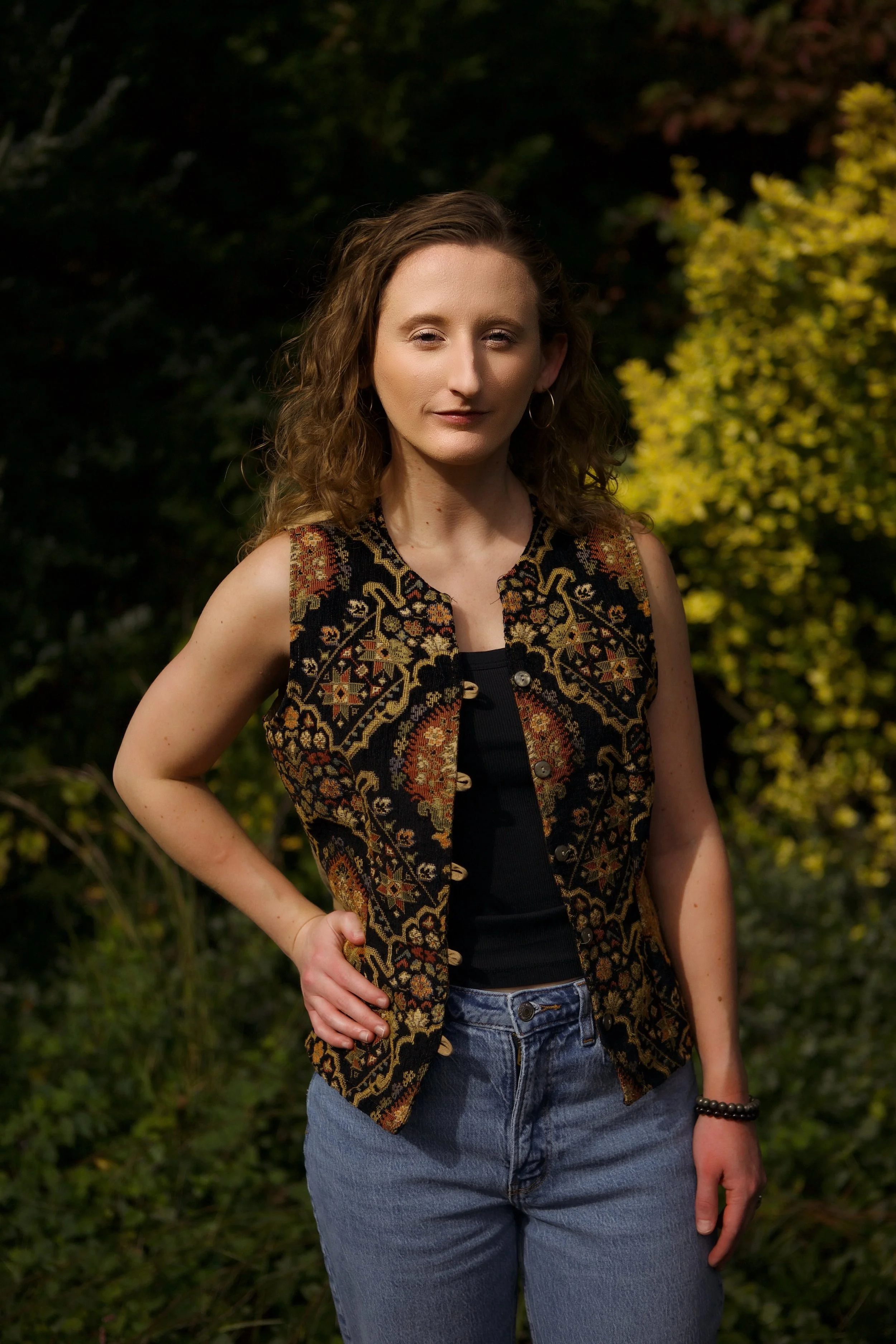 A woman with wavy brown hair standing outdoors in front of green and yellow foliage, wearing a sleeveless embroidered vest over a black top, blue jeans, and a black beaded bracelet.