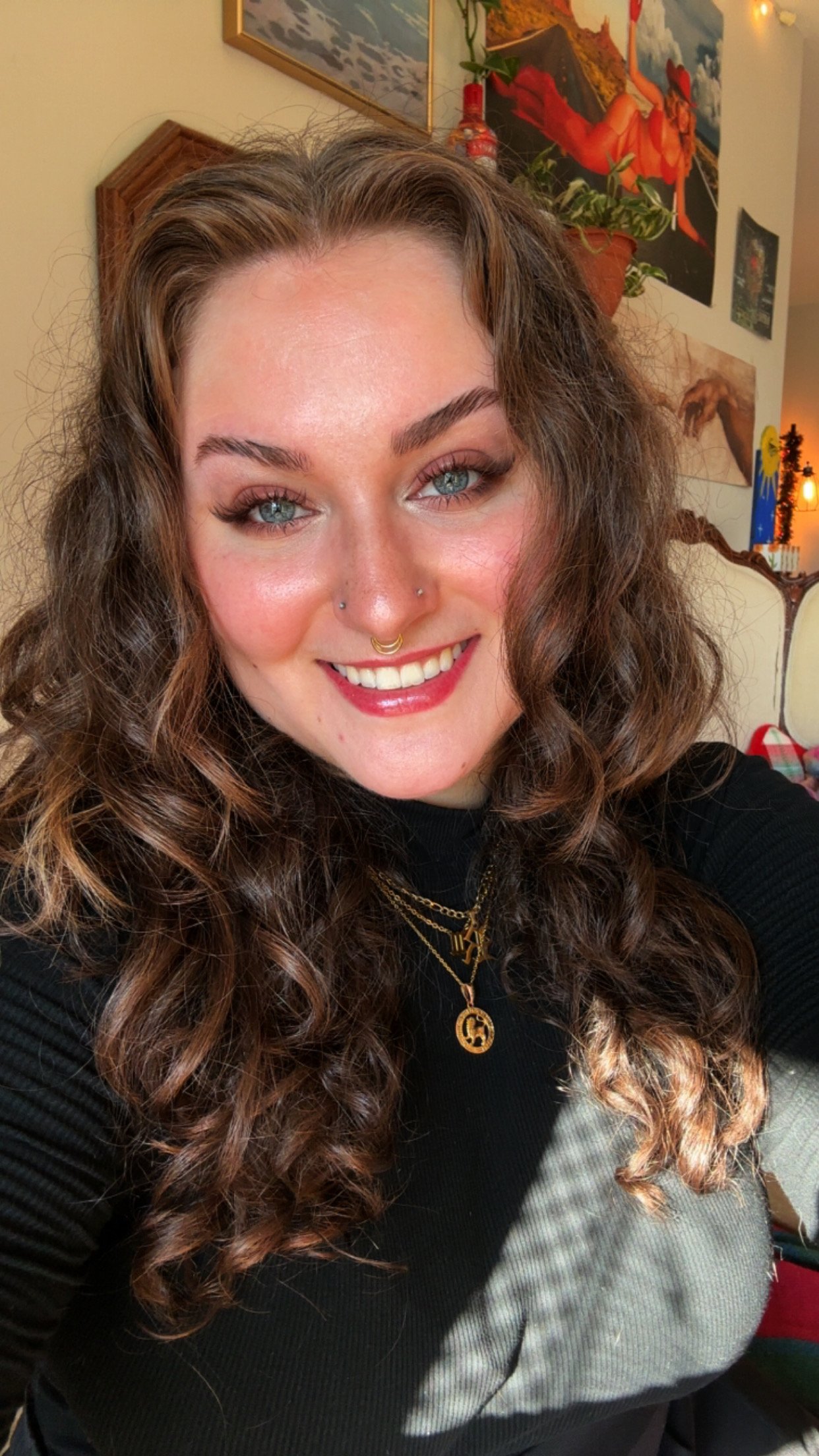 A woman with curly brown hair, blue eyes, and a septum piercing smiling in a room decorated with paintings, plants, and warm lighting.