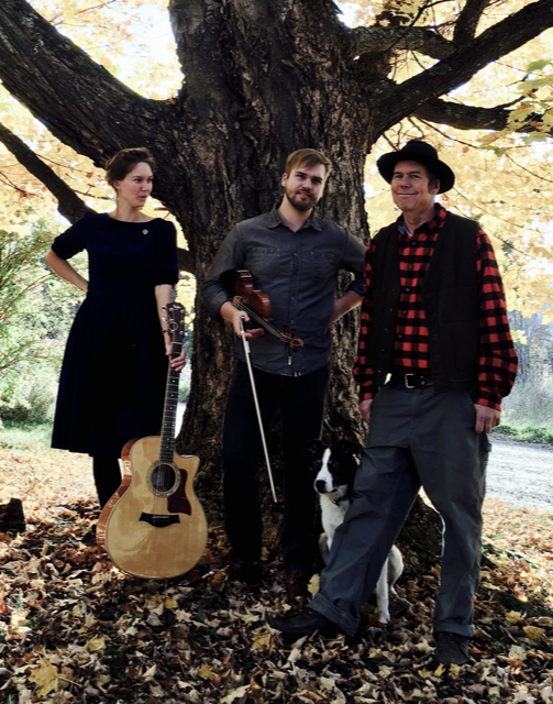 Black and white image of three musicians performing outdoors; one plays a violin, another plays a wooden flute, and the third plays an acoustic guitar.