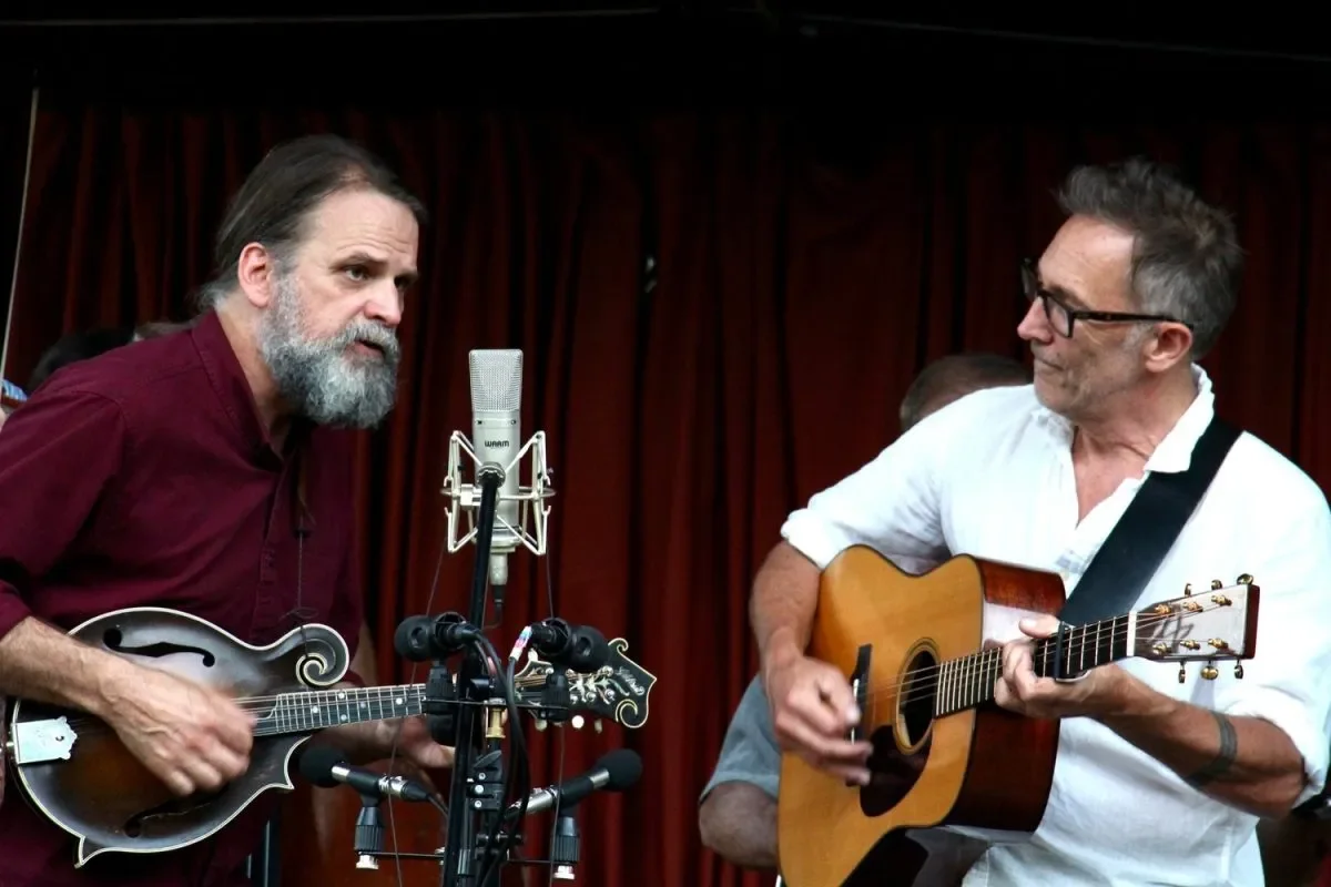 Two musicians performing on stage, one playing a mandolin and the other playing a guitar, with a microphone between them, in front of a red curtain.