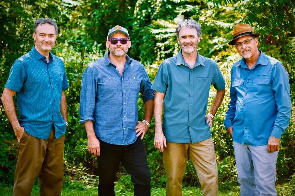 Four men standing on grass with trees and a hill in the background. The men are dressed in casual and semi-formal attire, including collared shirts and a vest and tie. It appears to be a daytime outdoor setting.